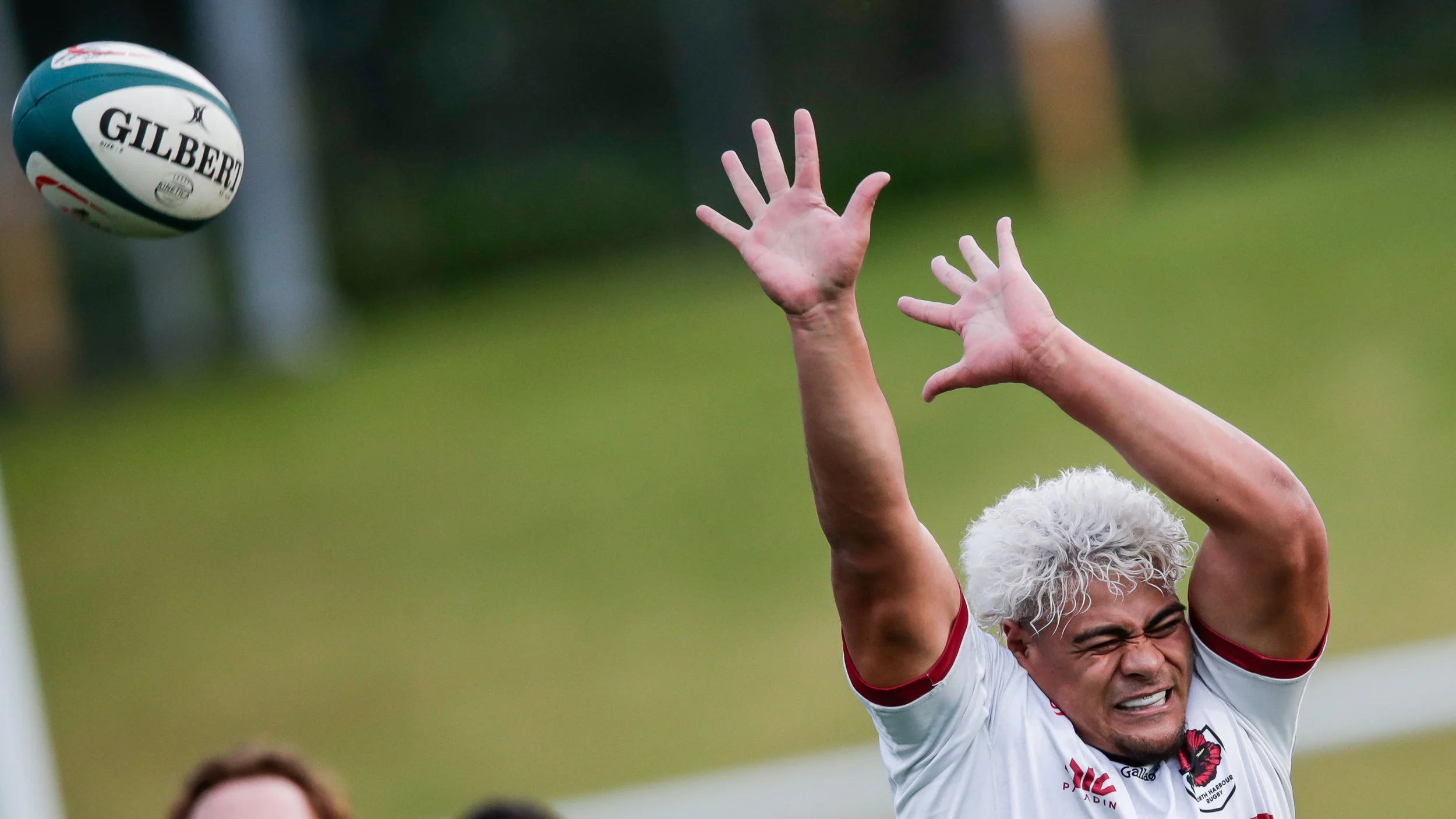 A rugby player with white hair and a white jersey, reaching to catch a rugby ball during a game.