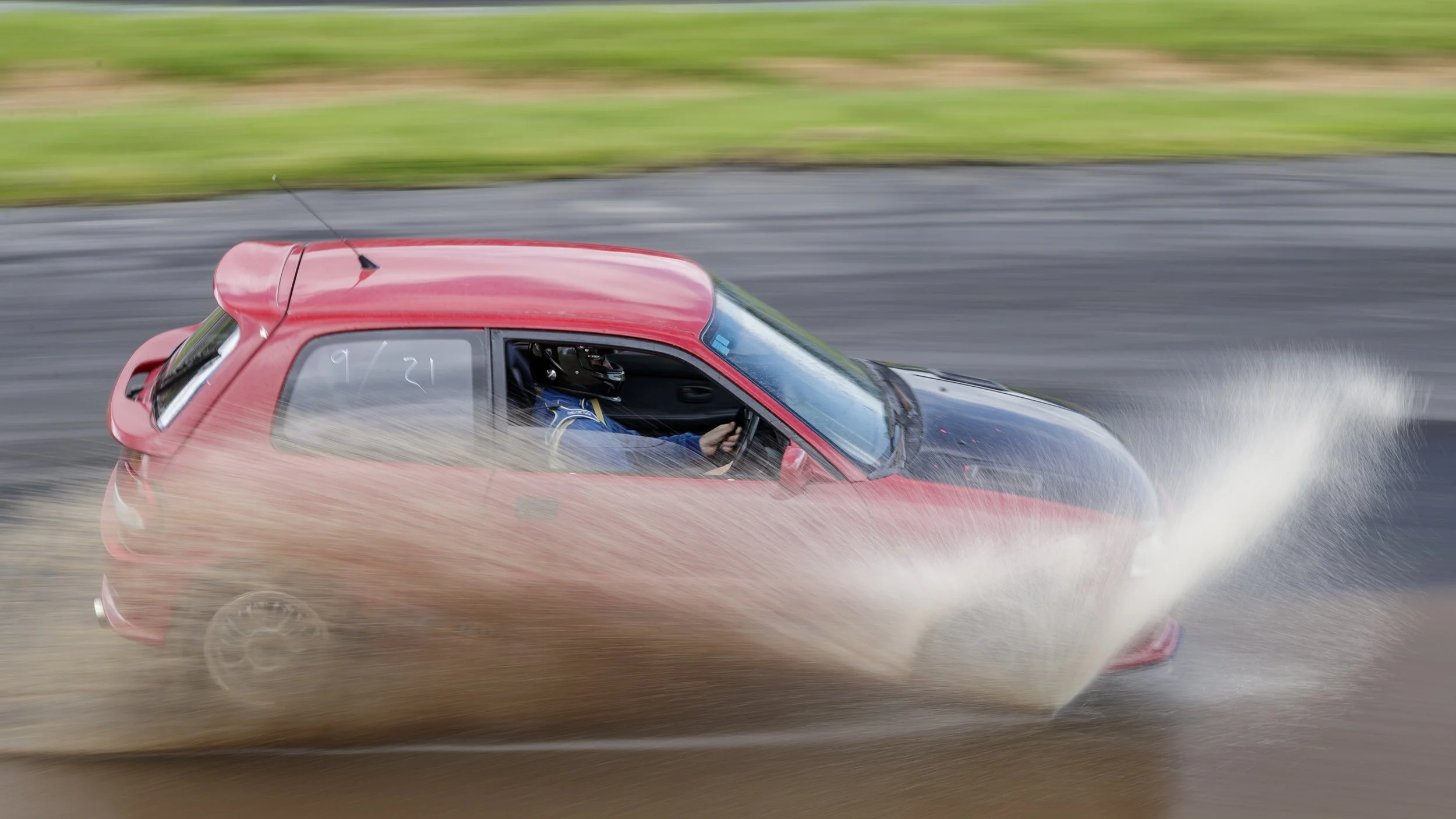 A red sports car racing on a track, kicking up dust, with a driver wearing a helmet visible through the window.
