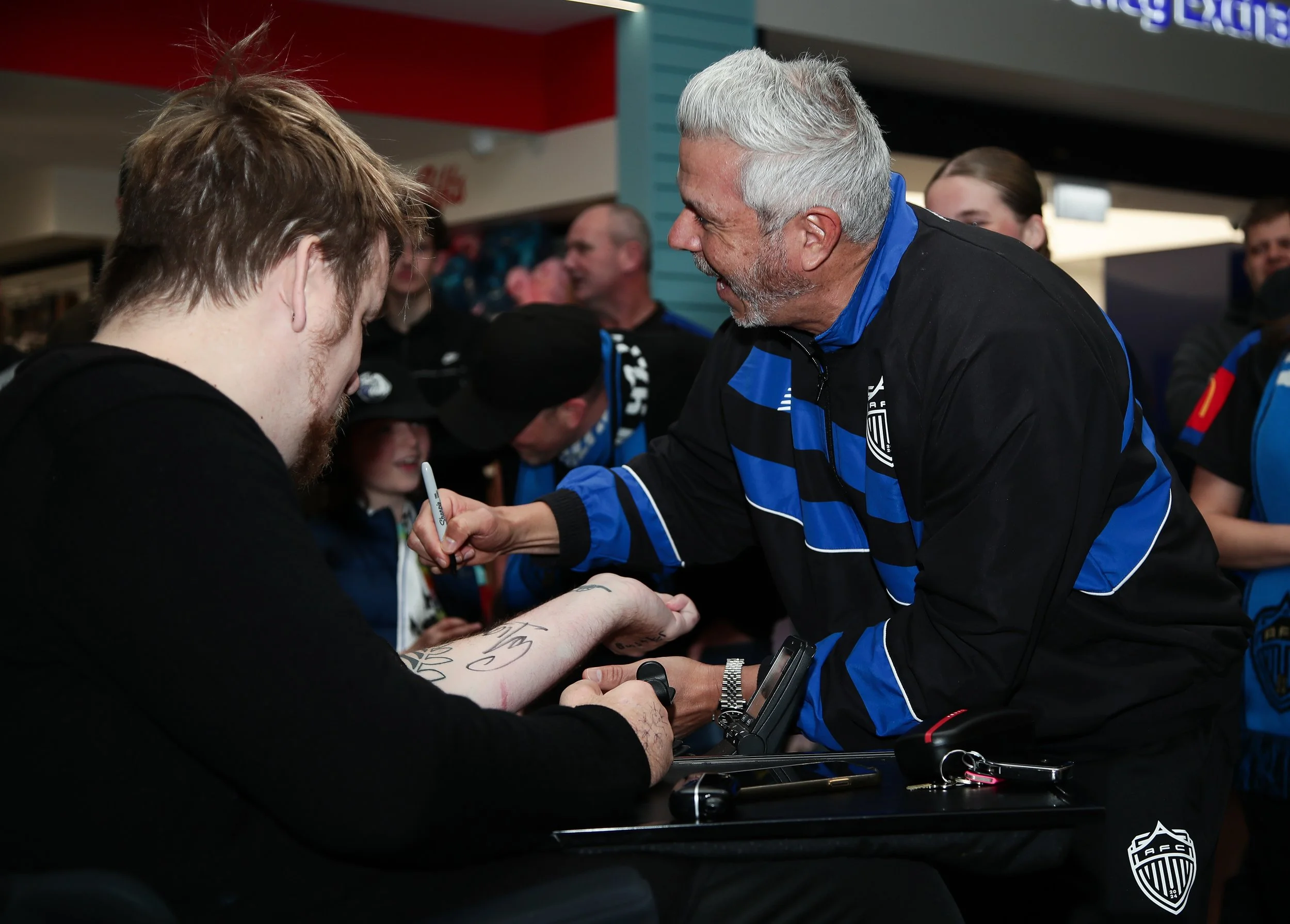 A man with gray hair and a beard wearing a black and blue jacket is signing an autograph for a younger man with red hair and a beard, who is holding a pen. Other people are gathered around, some smiling, in an indoor setting, possibly at a fan event 