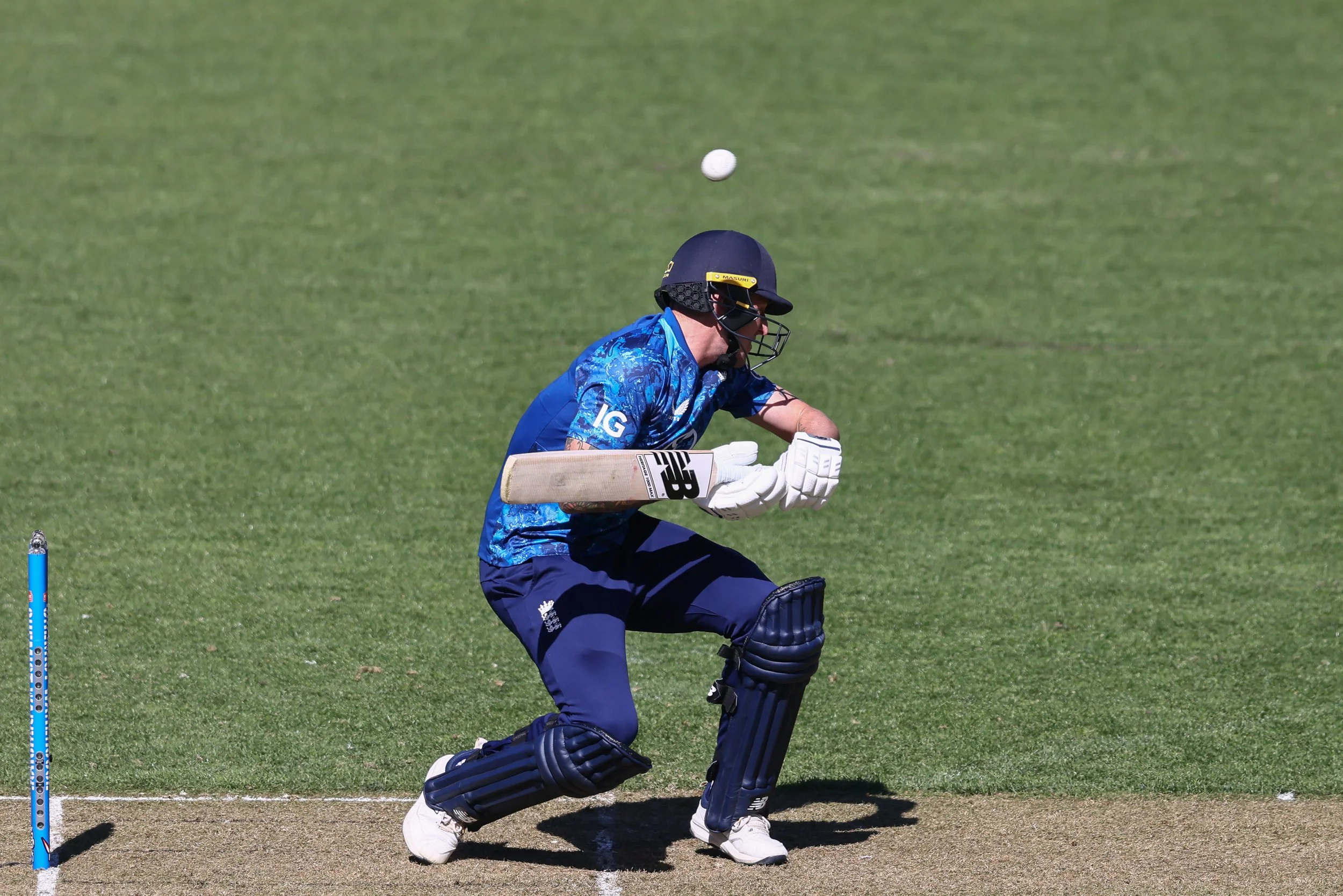 A cricket player in blue uniform and helmet, wearing gloves, is ducking on the cricket pitch with a ball in the air.