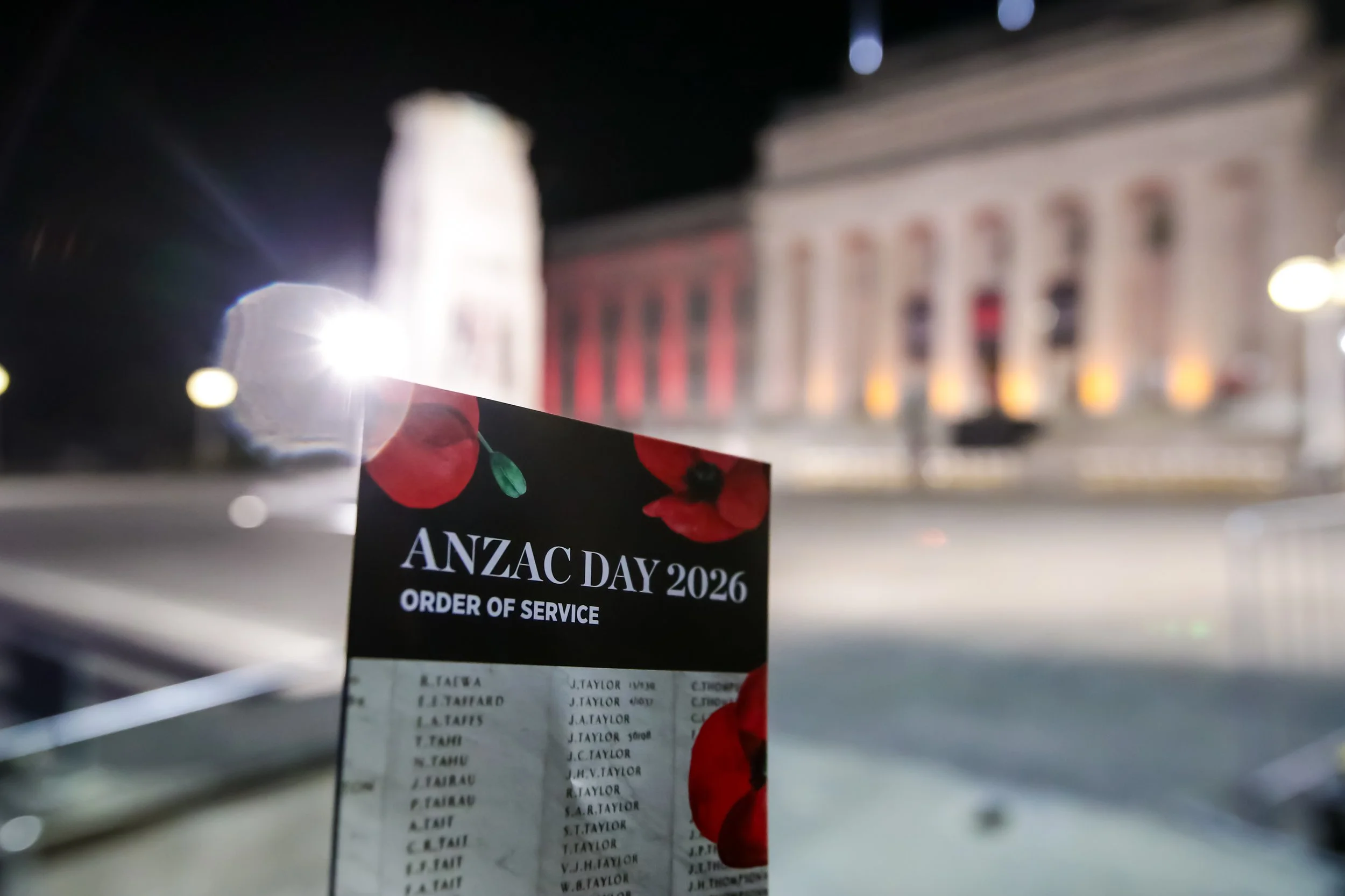 A close-up of a banner or sign for ANZAC Day 2026 resting on an outdoor surface with a blurred urban background at night.