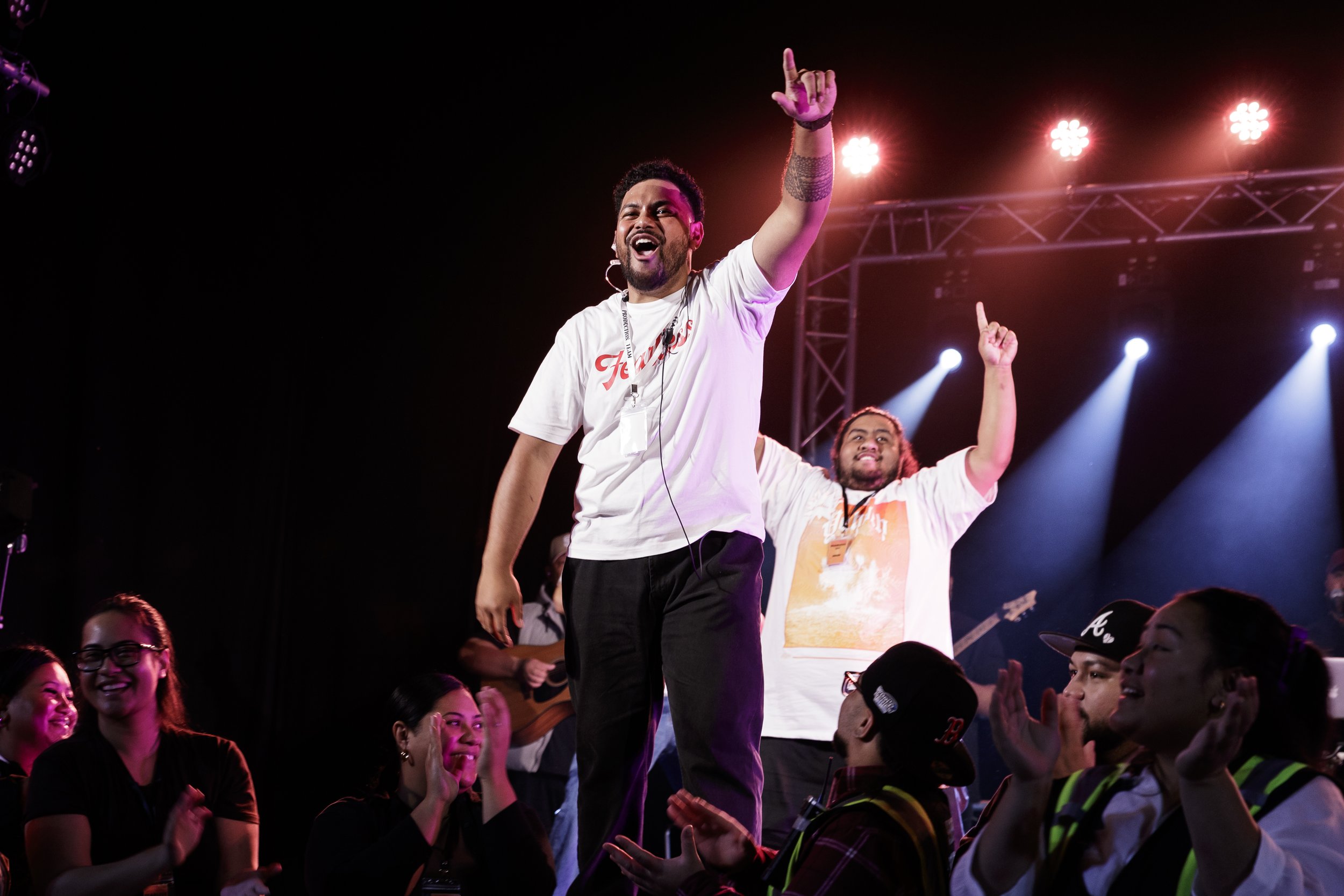 A man with curly hair and facial hair, wearing a white T-shirt with red lettering, stands on a stage with one arm raised pointing upwards, smiling. The stage has bright lights and a crowd of diverse people cheering and clapping around him.