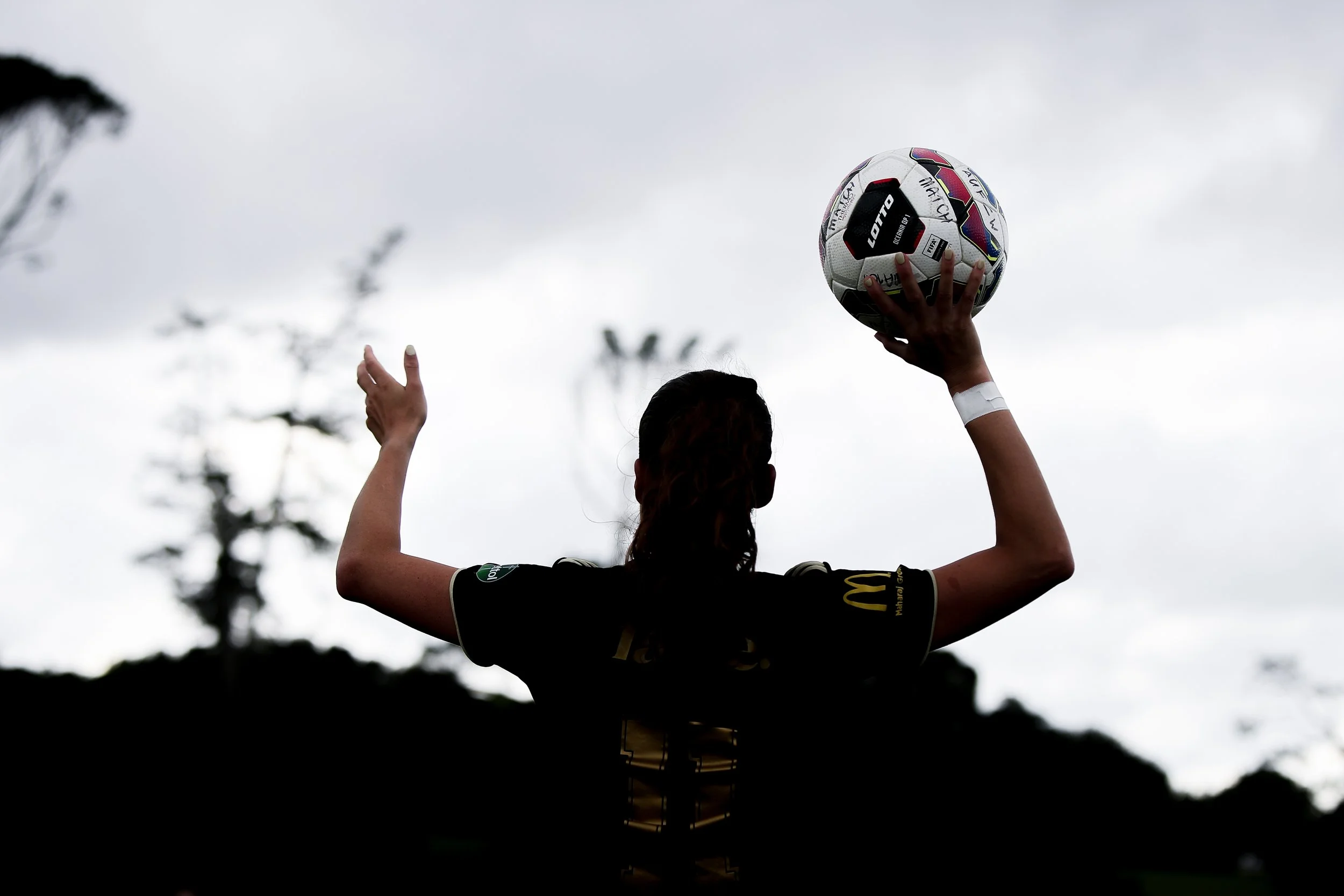 A person in a black sports jersey holding a soccer ball above their head outdoors, with trees and cloudy sky in the background.