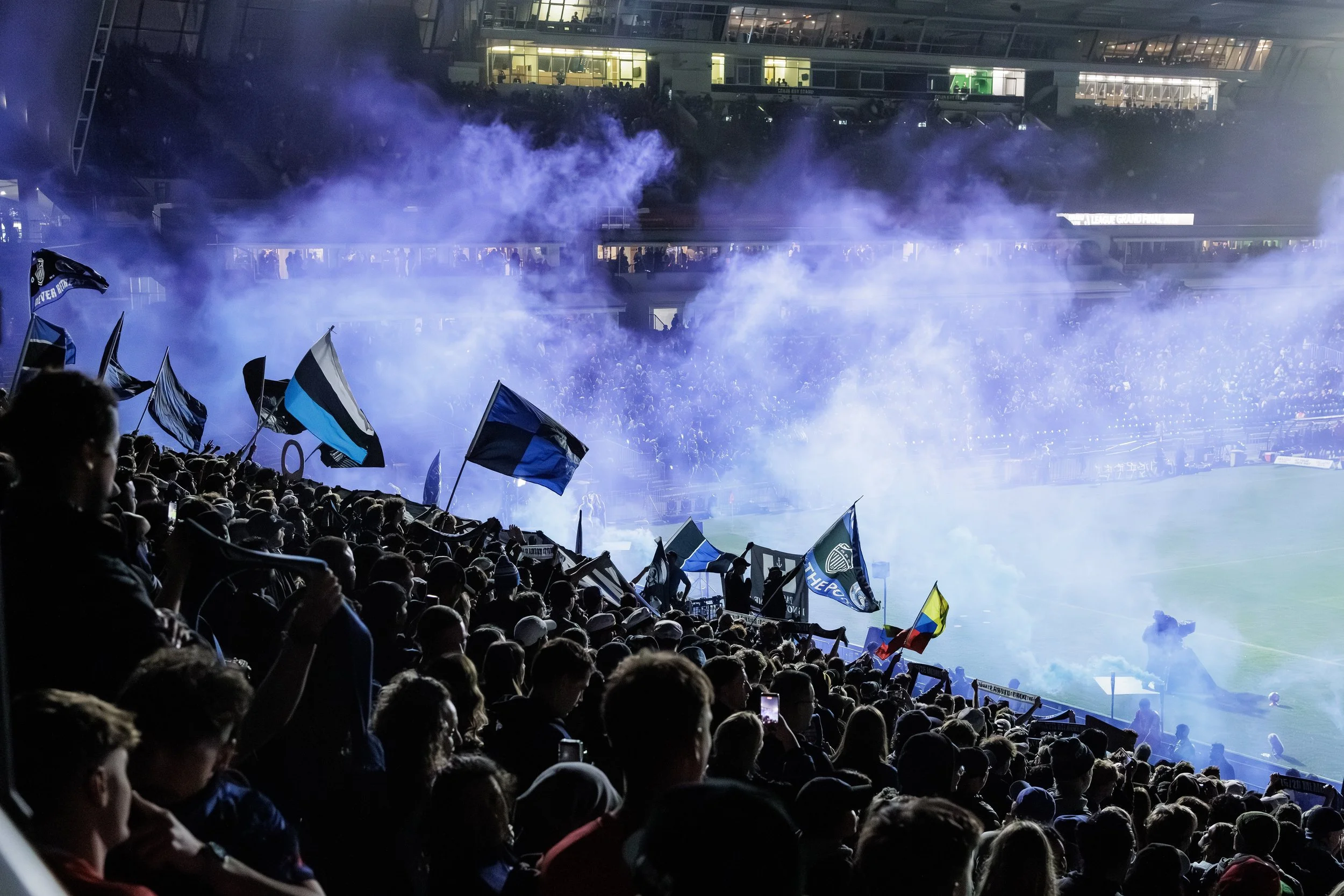 Crowd of soccer fans in a stadium with flags and smoke, watching a game at night.