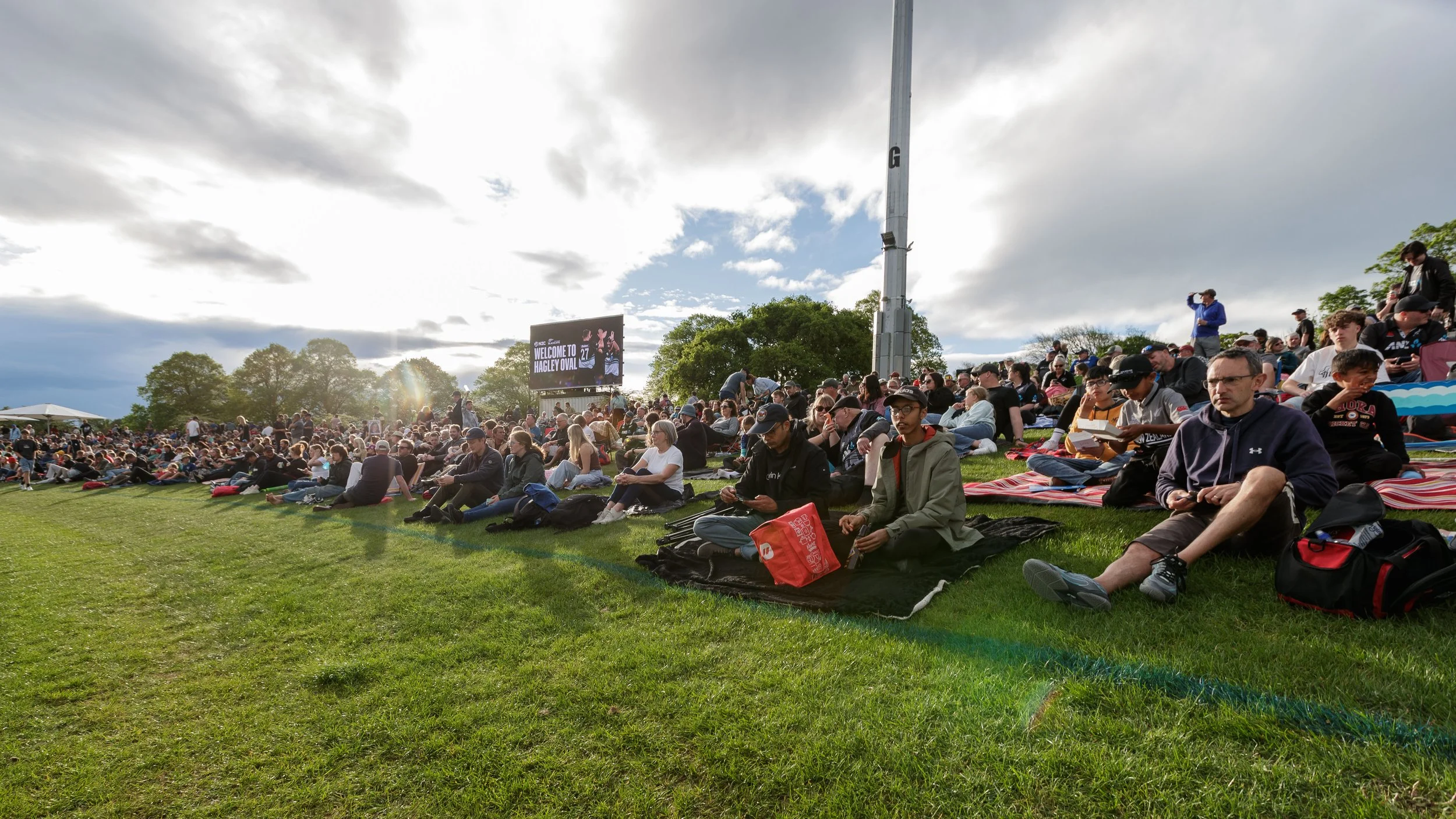Crowd of people sitting on the grass at an outdoor event near Hagley Oval, with a large scoreboard, trees, and cloudy sky in the background.