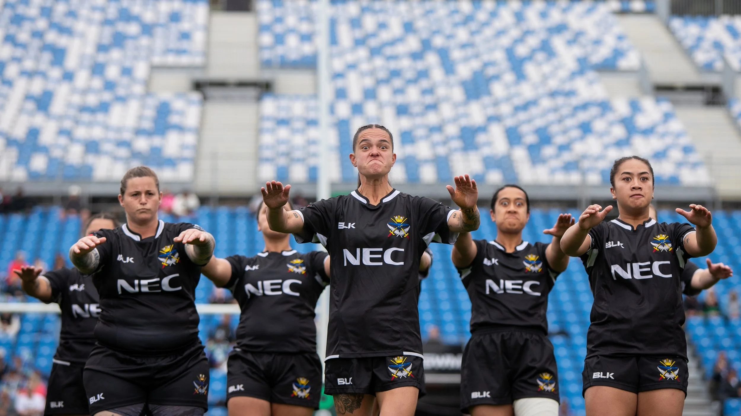 Young women in black sports uniforms performing a haka on a sports field with stadium seating in the background.