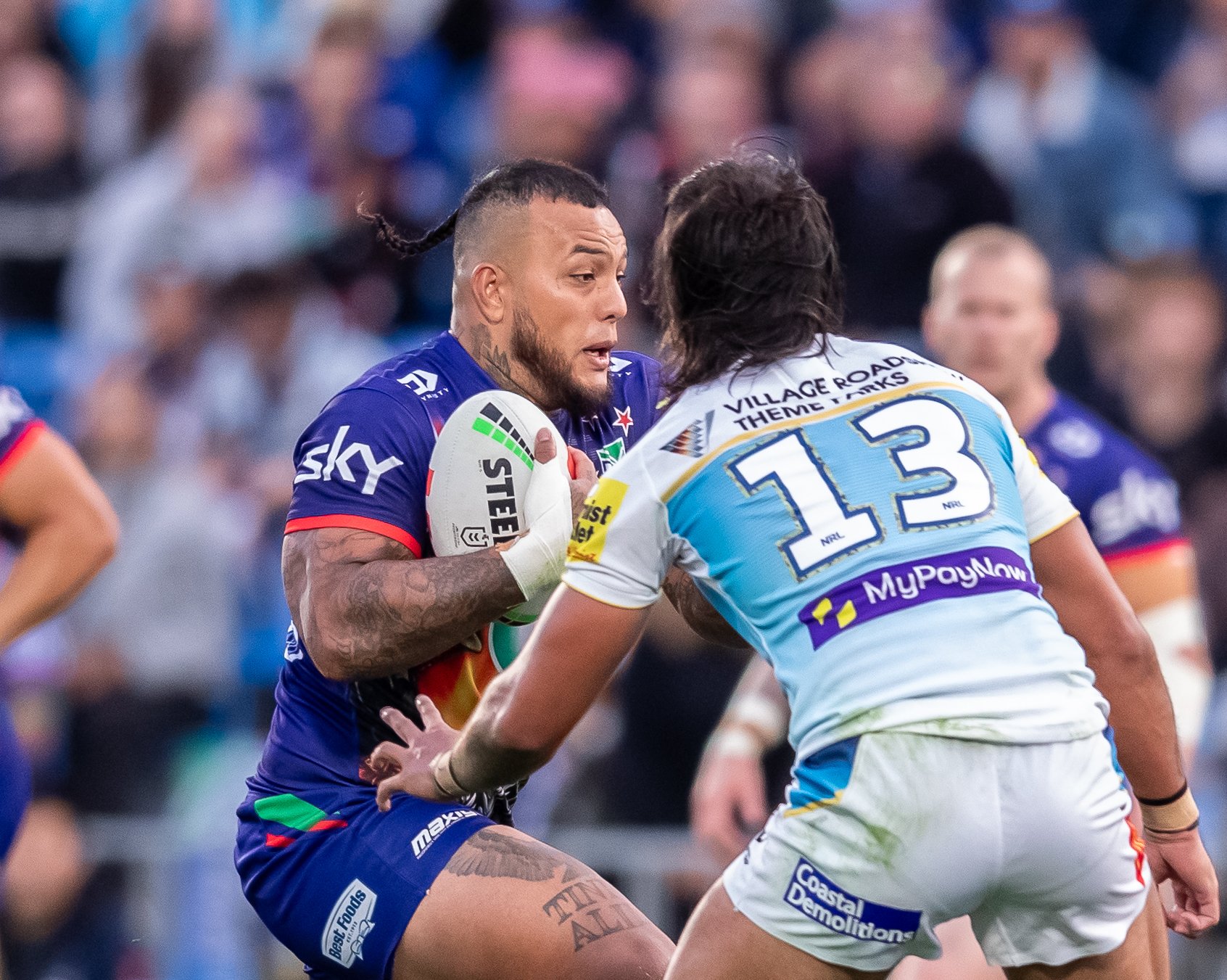 Two rugby players in an intense moment during a game. One player, wearing a blue jersey, holds a rugby ball, and the other, in a white jersey with the number 13 on her back, is approaching him. The background shows blurred spectators in the stands.