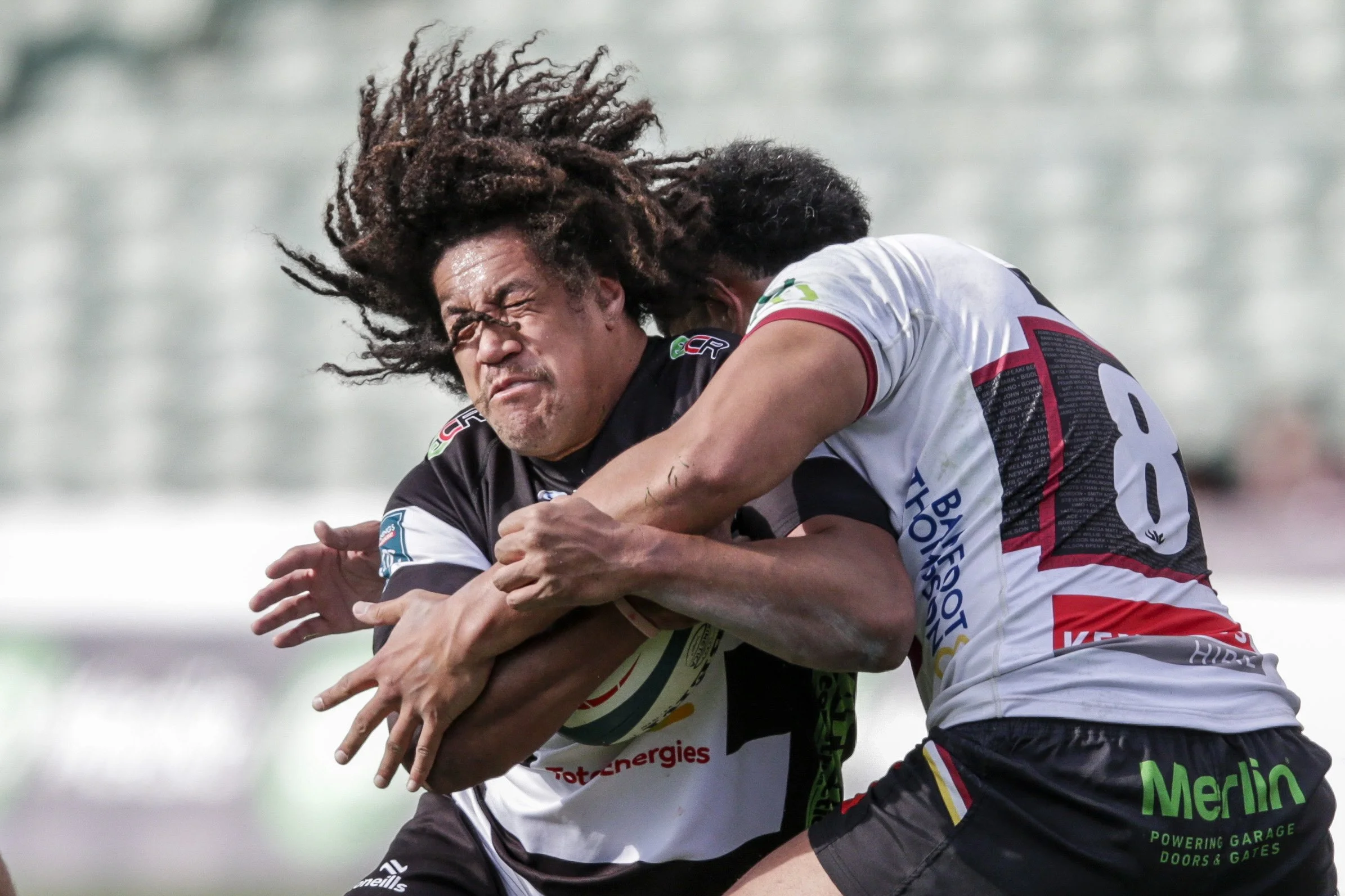Two rugby players are engaged in a tackle during a game, with one player holding onto the other tightly. Both players are wearing game jerseys, and their hair is flying as they struggle for control.