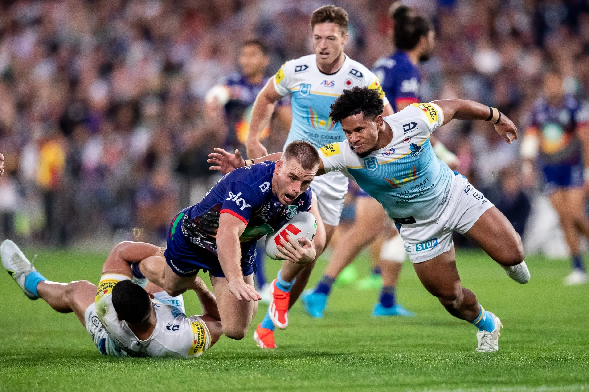 A rugby players is tackled during a game with a blurred crowd in the background.