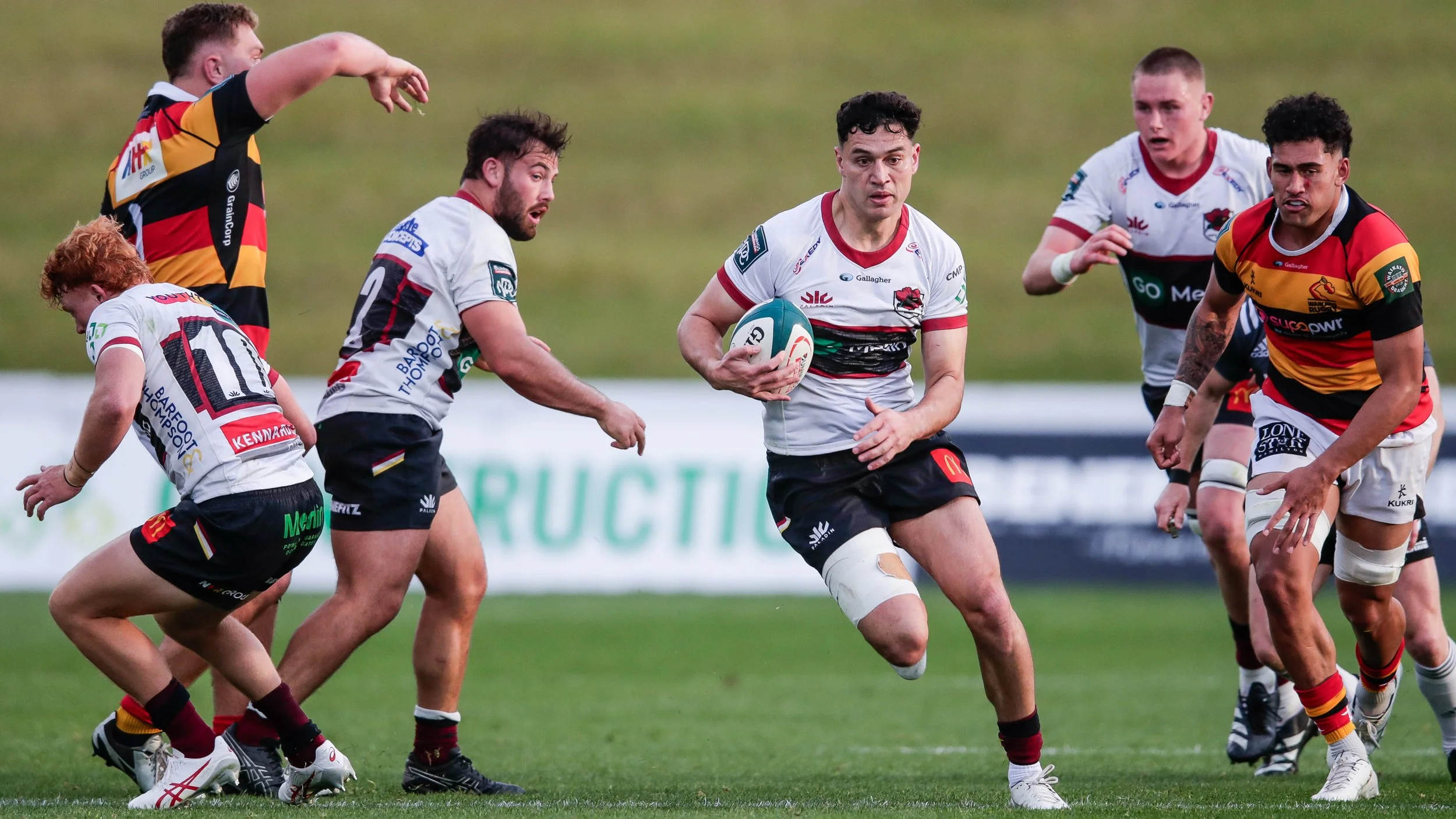 Rugby players in action during a game, with one player holding the ball and others moving around him on the field.