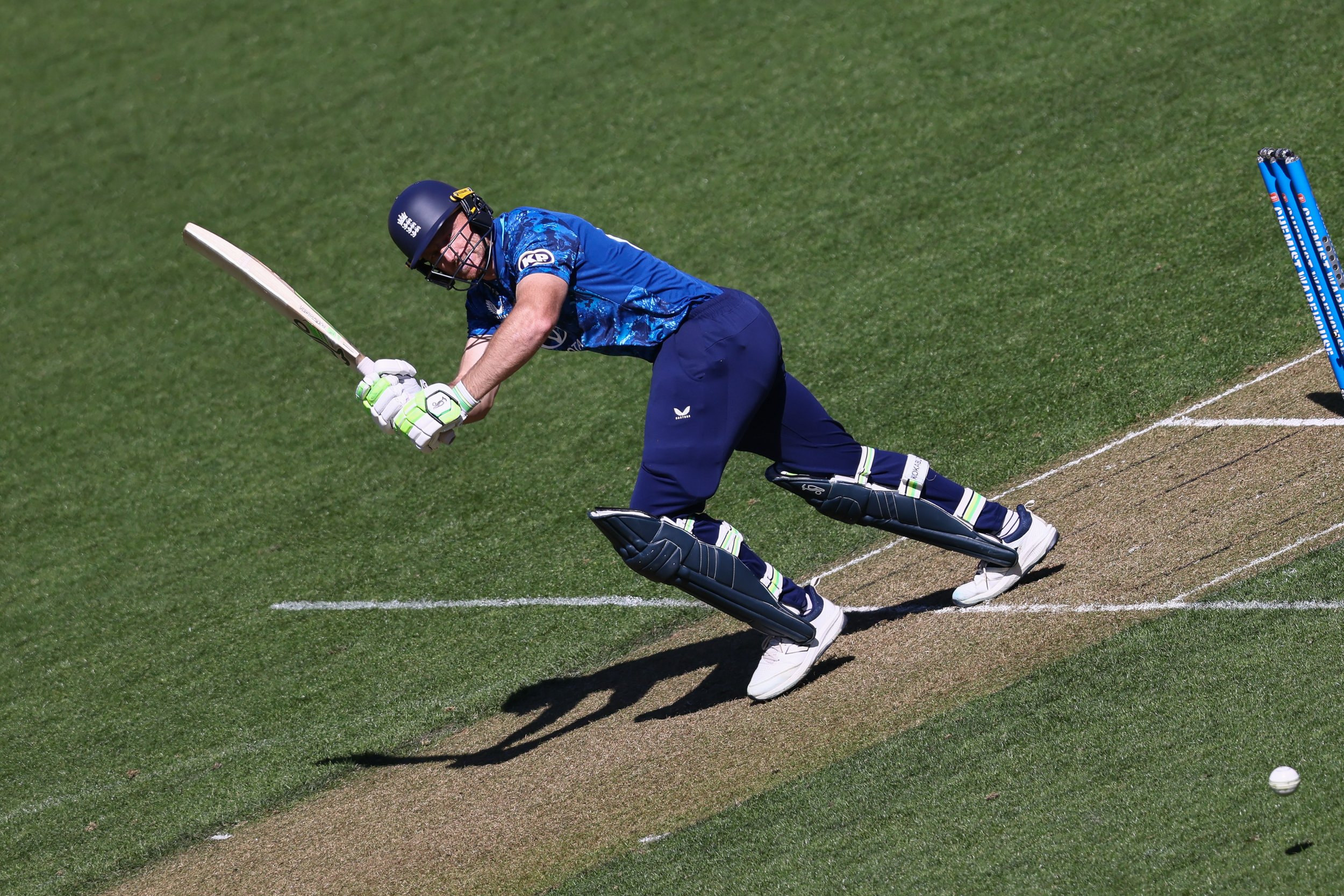 A cricket player in blue uniform and helmet is sliding on the pitch to reach the cricket ball.