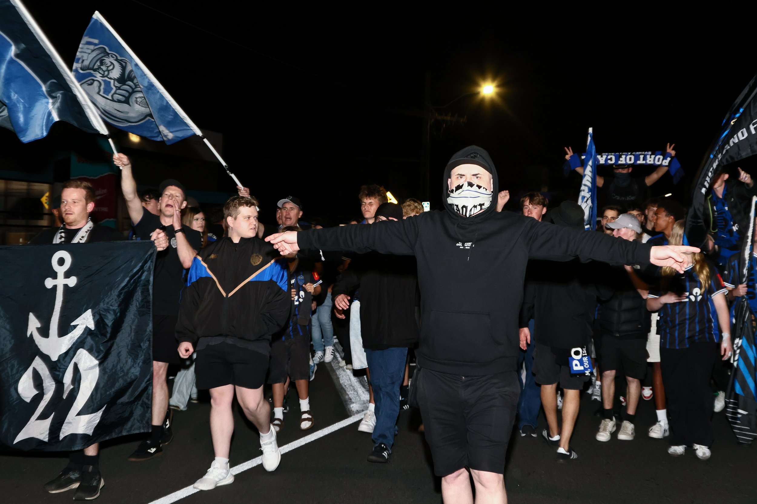 A group of people participating in a nighttime march, many holding flags and banners, with a man in the foreground wearing a black hoodie and a bandana covering his face.