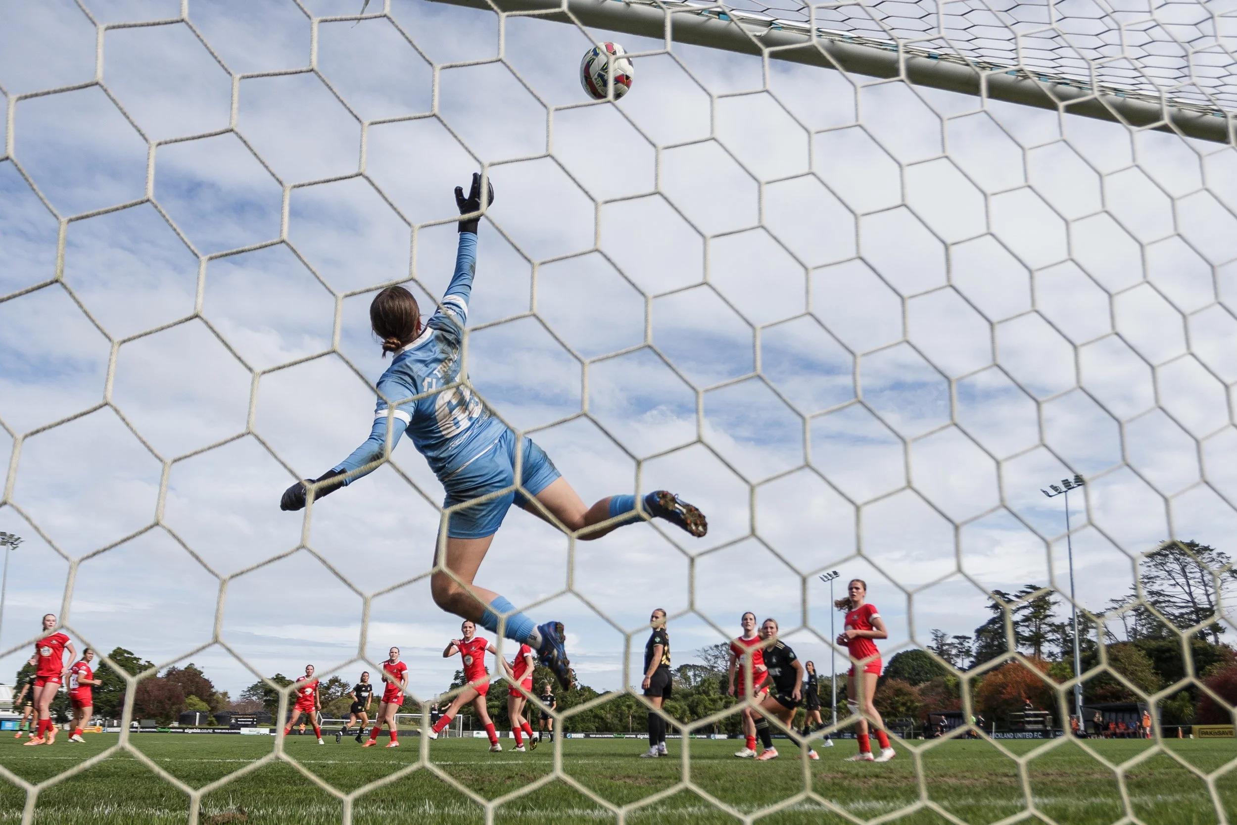 A female soccer goalkeeper in a blue uniform reaching up to block a ball from entering the goal during a match, with other players and spectators visible on the field.