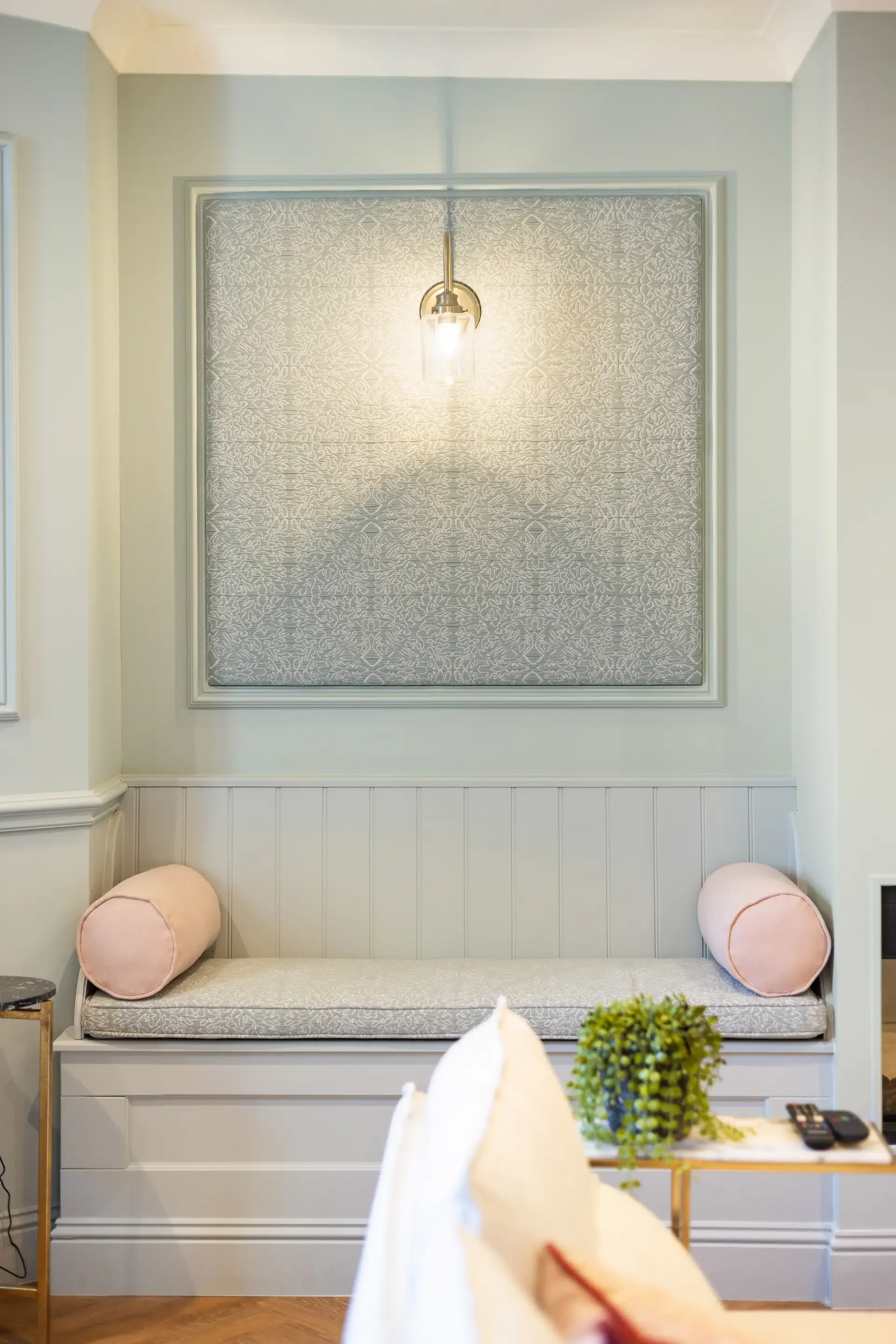 A light-colored sofa with two pink cushions, a small side table with a round top and gold legs, and a decorative wall panel with intricate embossed pattern. A hanging light fixture and some greenery on a table are visible in the scene.