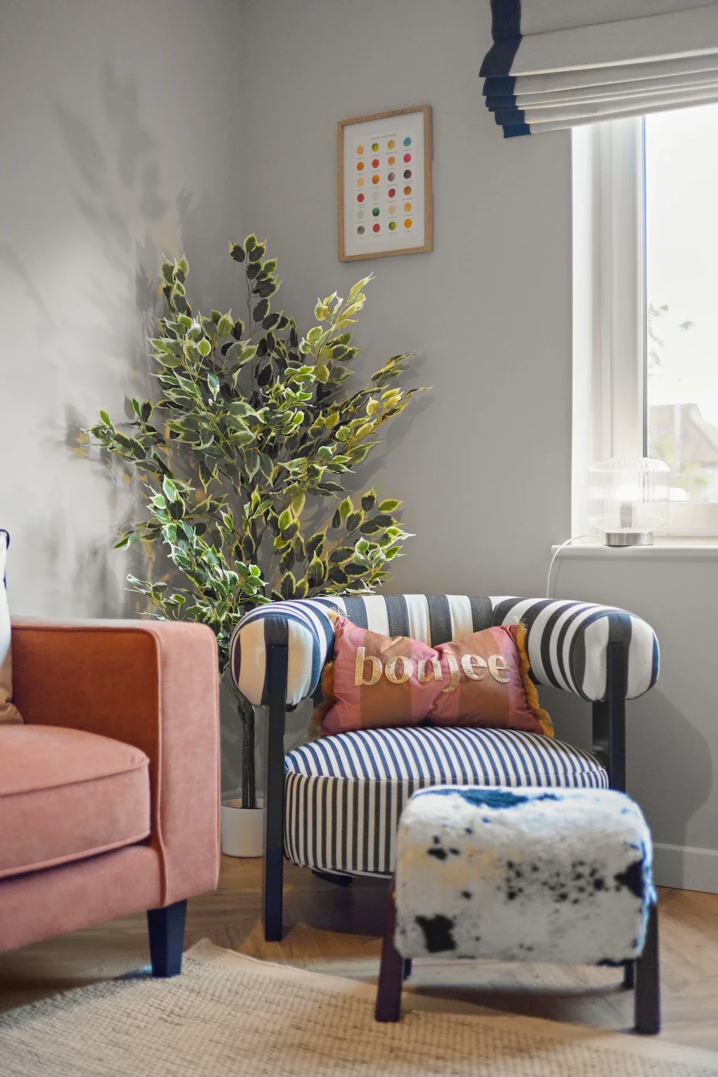 A cozy living room corner with a striped armchair, a pink pillow with the word 'boujee,' an upholstered footstool with a black and white cow print, a pink velvet sofa, a large leafy plant, a framed print of colorful dots on the wall, and a window with blinds letting in natural light.