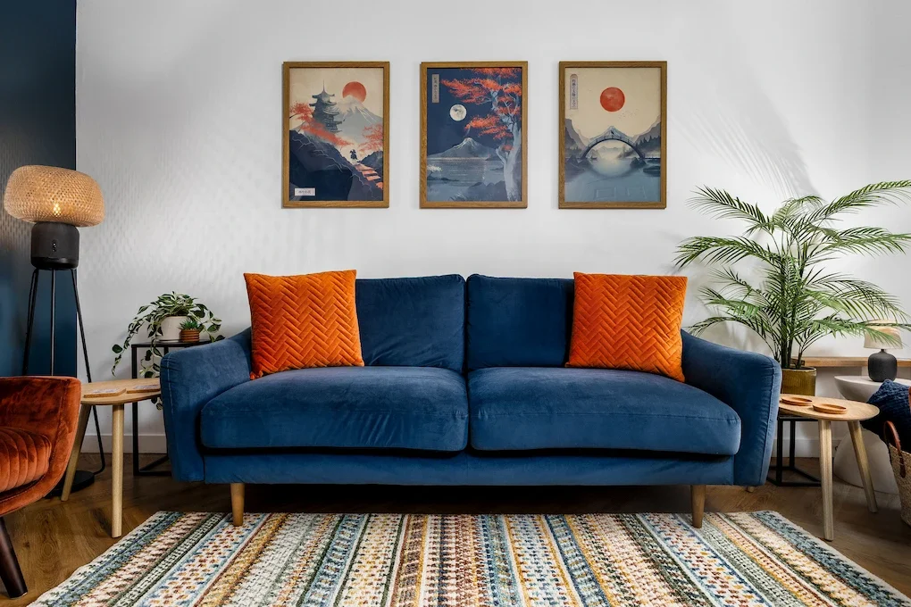 A living room with a blue sofa and two orange pillows, a patterned area rug, a side table with a plant, a wooden coffee table, and framed Asian landscape artwork on the white wall.