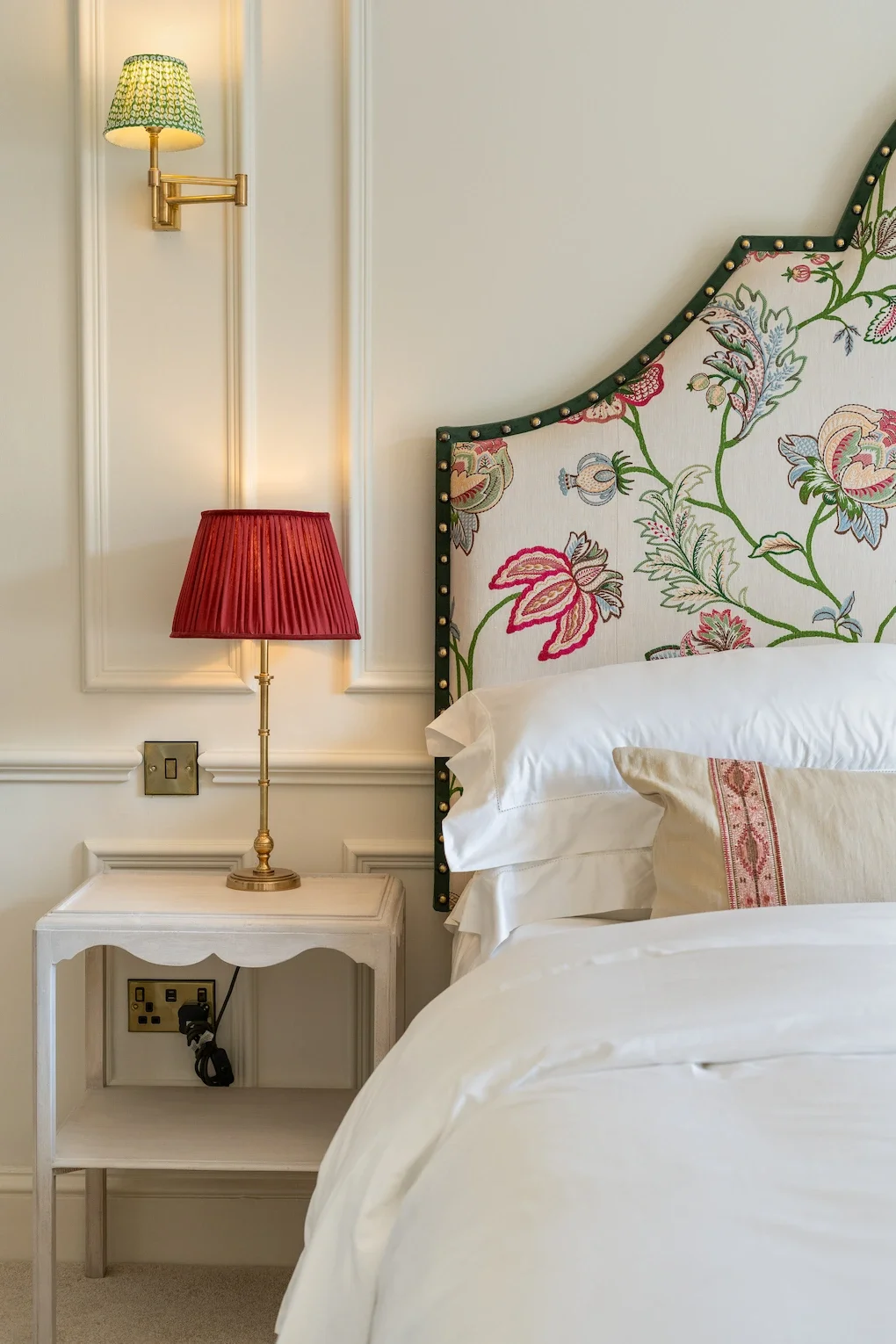 A bedroom corner featuring a floral-patterned headboard with a dark green border, a white nightstand with a brass table lamp with a red lampshade, a wall-mounted yellow-green lampshade light, and white bedding with a decorative pillow.