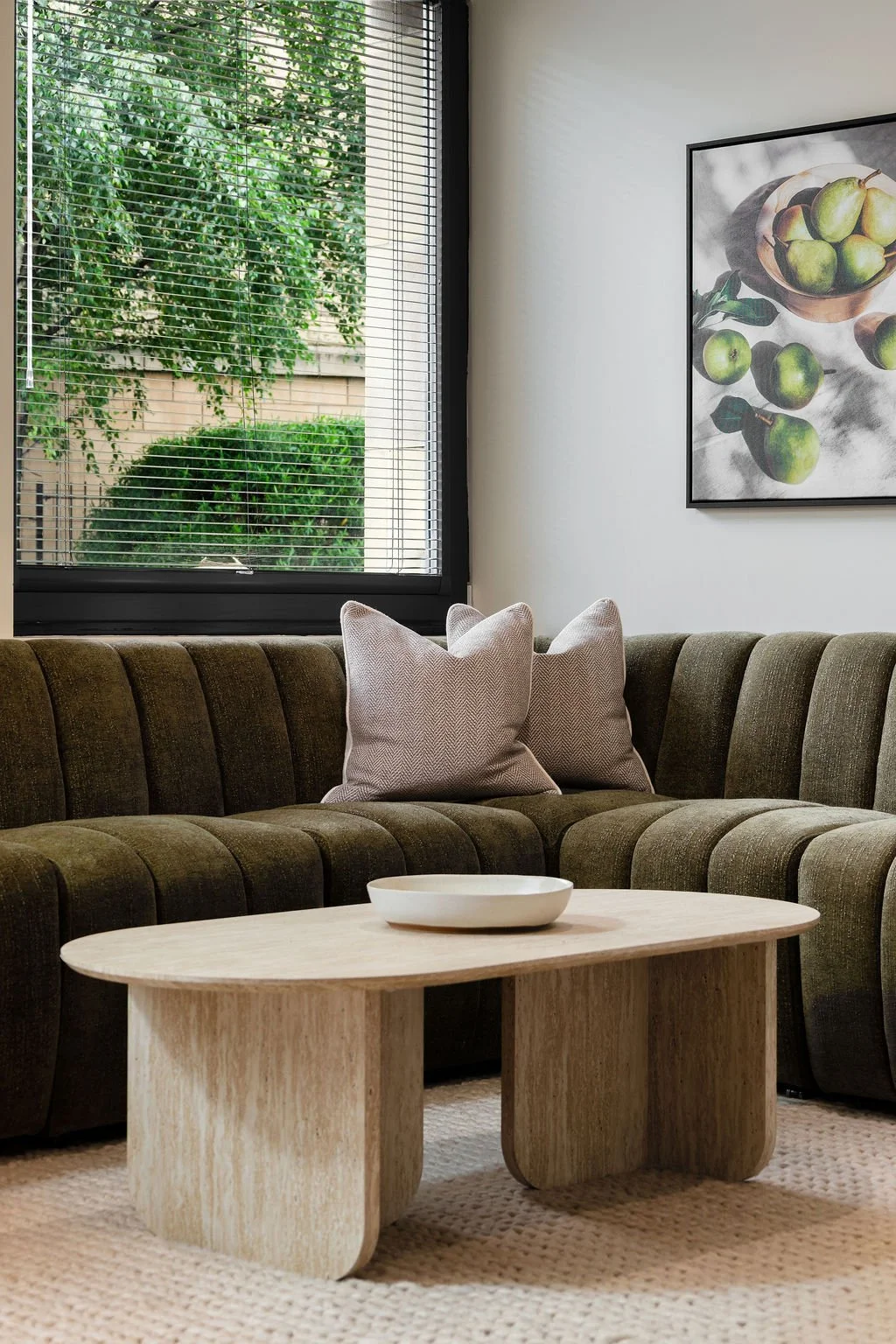 Living room with a green couch, two gray pillows, a wooden coffee table with a white bowl, a large window with blinds revealing green foliage outside, and a framed art print of green apples on the wall.