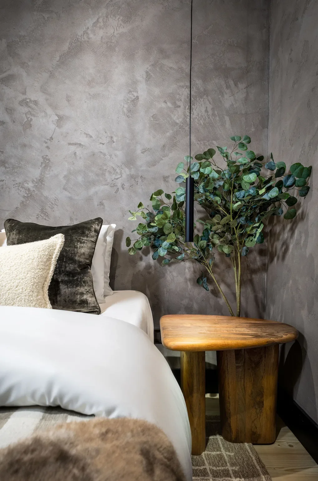 A cozy bedroom corner featuring a bed with dark and light-colored pillows, a wooden side table, a tall green leafy plant, and a textured gray wall with a black vertical pendant light.