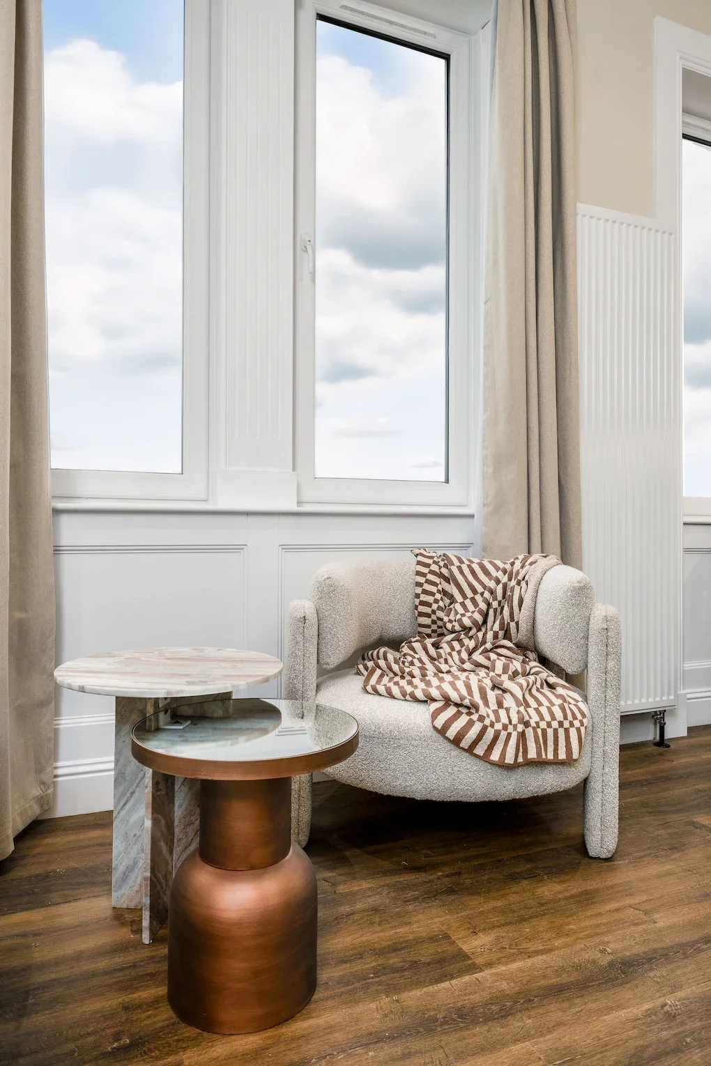A cozy living room corner with a white textured armchair draped with a brown and beige checkered blanket. Two round side tables, one with a marble top and the other with a metallic finish, are placed next to the armchair. Large windows with beige curtains allow natural light into the space.