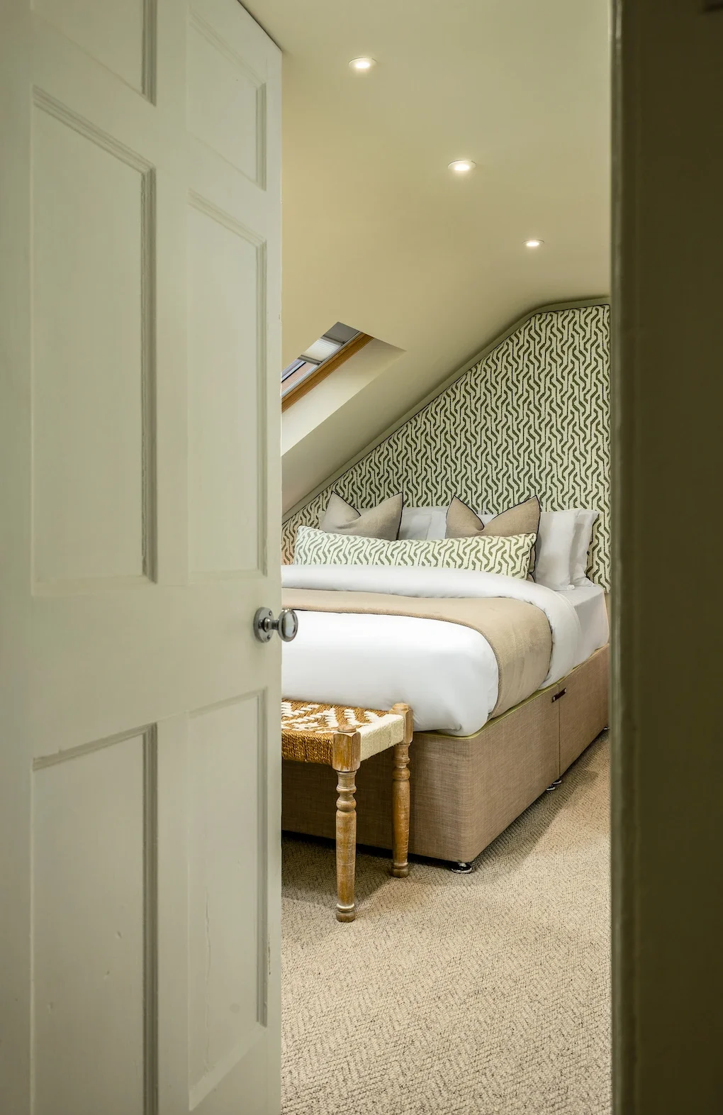 A cozy bedroom with a sloped ceiling, a skylight window, decorative pillows, and a patterned headboard, viewed from behind a partially opened door.