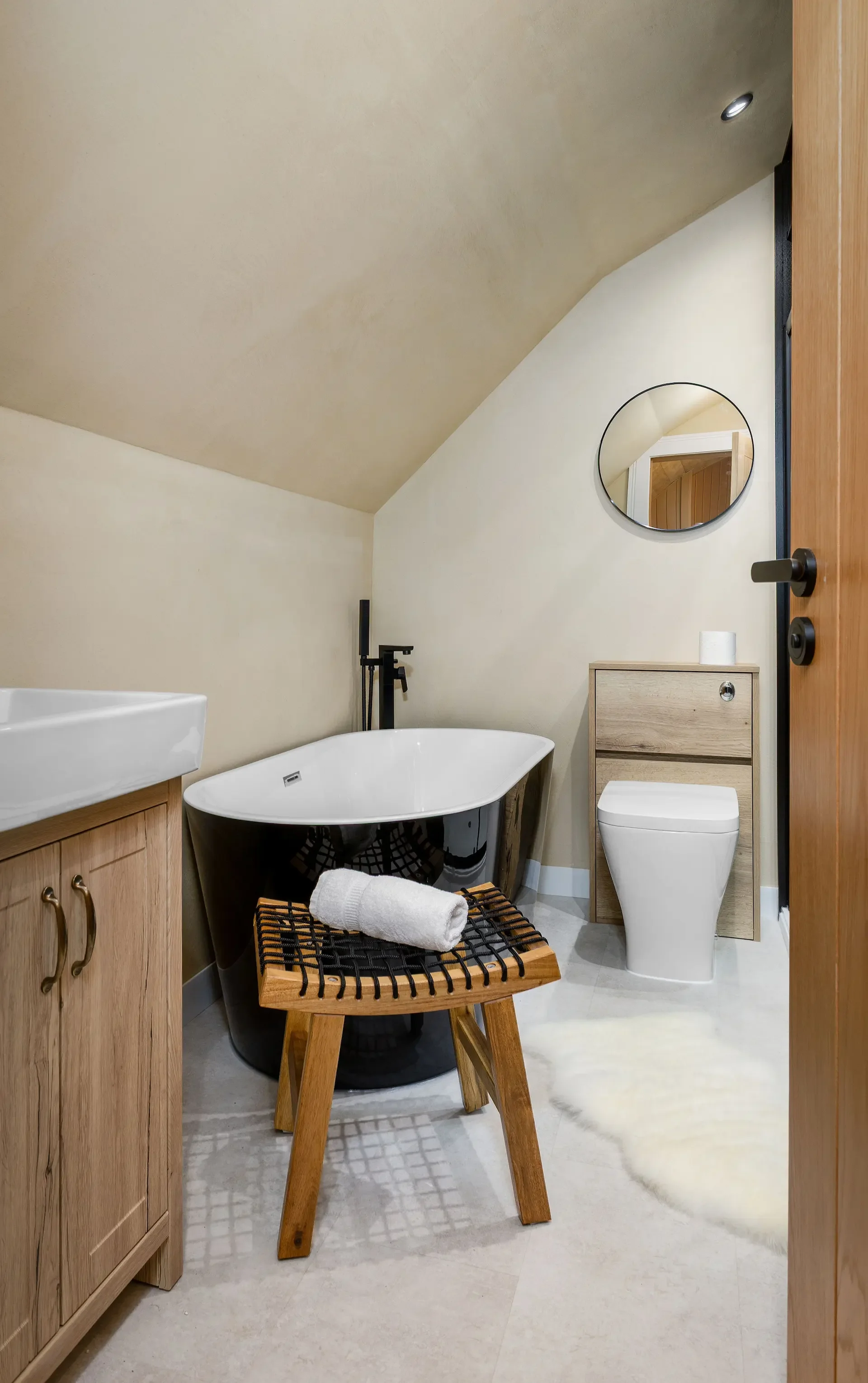Modern bathroom featuring a black bathtub, a small wooden stool with a rolled towel, a vanity with a white sink, a toilet, a round mirror, a small cabinet with a roll of toilet paper, a wooden door, and wood accents, with beige walls and flooring.