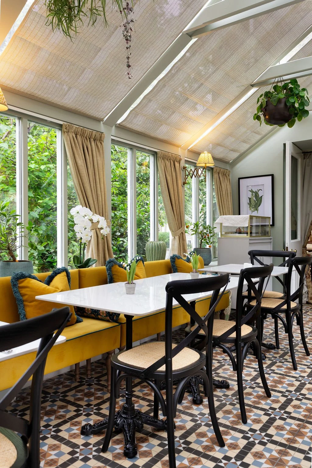 Bright dining room with large windows, beige curtains, yellow cushioned bench, black chairs, potted plants, framed artwork, patterned tile floor, and ceiling with skylights and hanging plants.