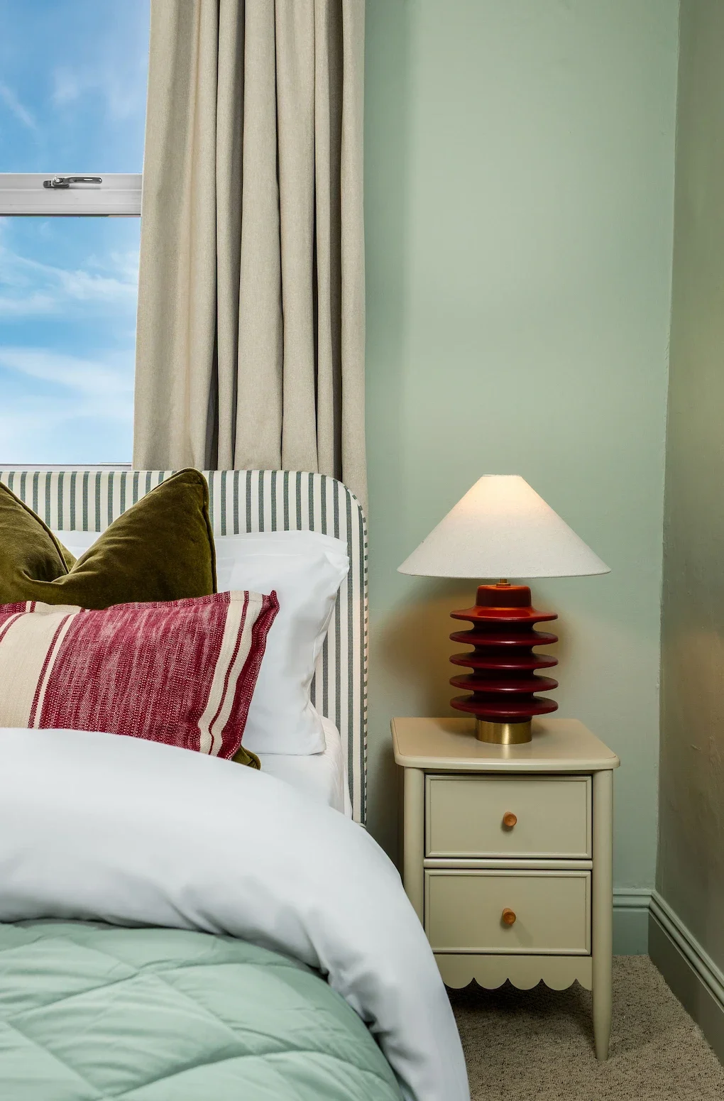 A cozy bedroom with a bed featuring striped and solid pillows, a nightstand with a modern red ceramic table lamp, and a window with beige curtains showing a partly cloudy sky.