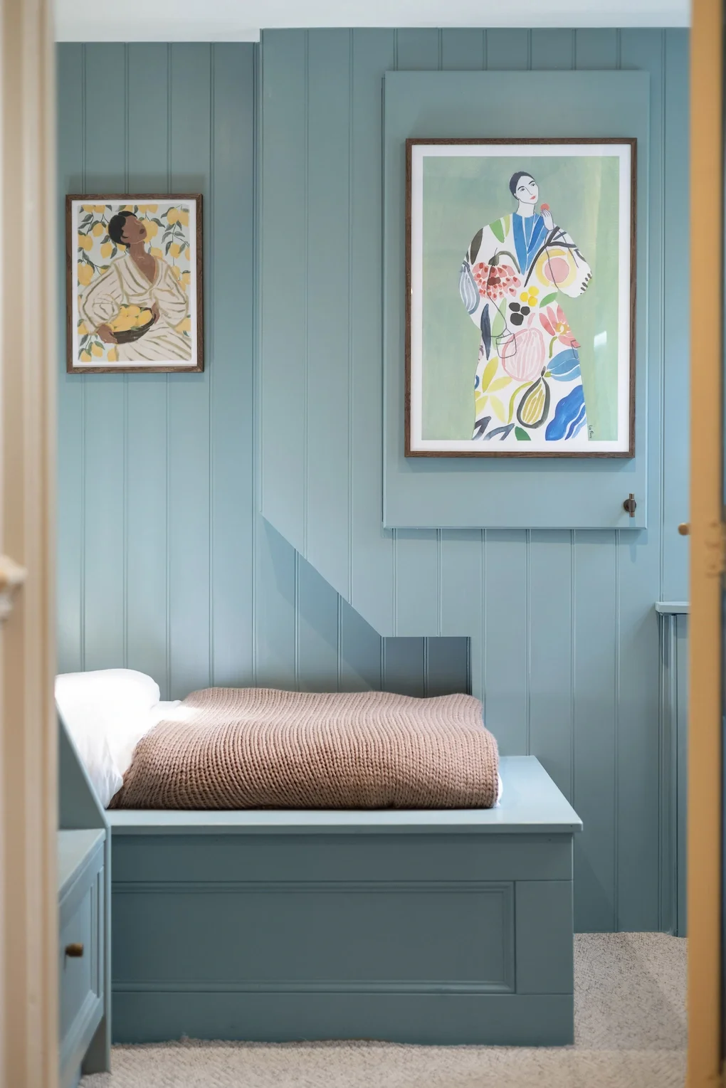 A cozy bedroom with light blue wooden panel walls, a neatly made bed with a white frame, white pillow, and a brown knitted blanket, two colorful modern art paintings hanging on the wall.