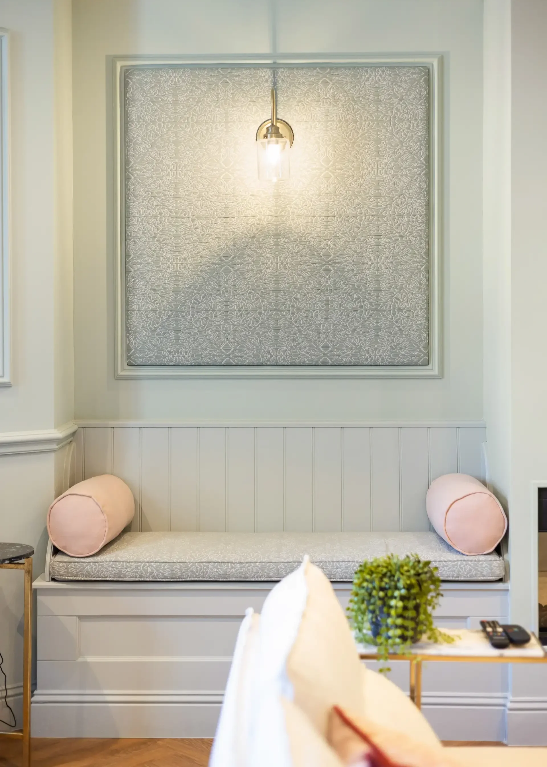 A bench with a patterned cushion, two pink round pillows, a small side table with a plant and remote controls, and a decorative wall with a light fixture above.