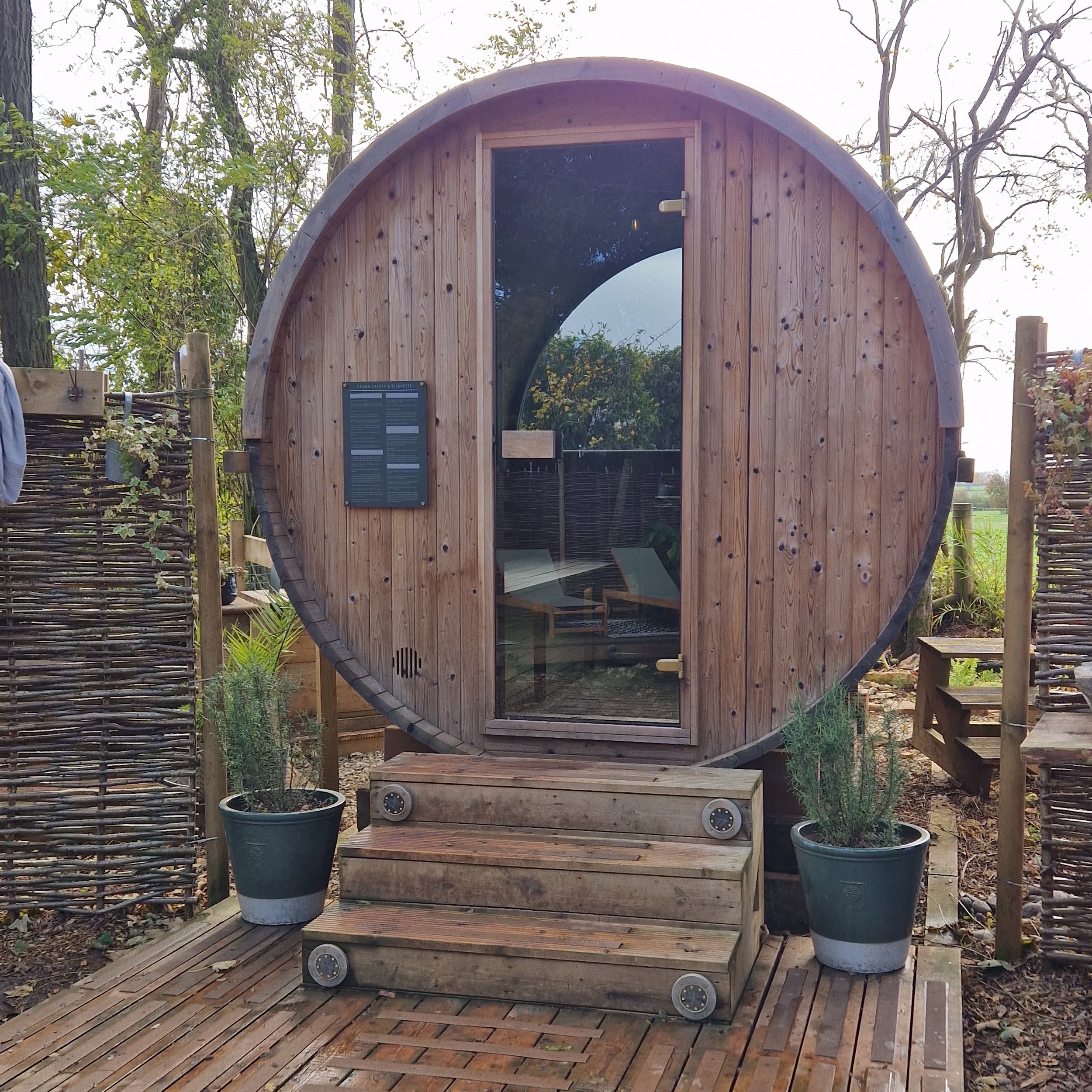 A small wooden sauna with a curved roof, glass door, and wheels at the bottom, placed on a wooden platform in a natural outdoor setting with trees and fencing.