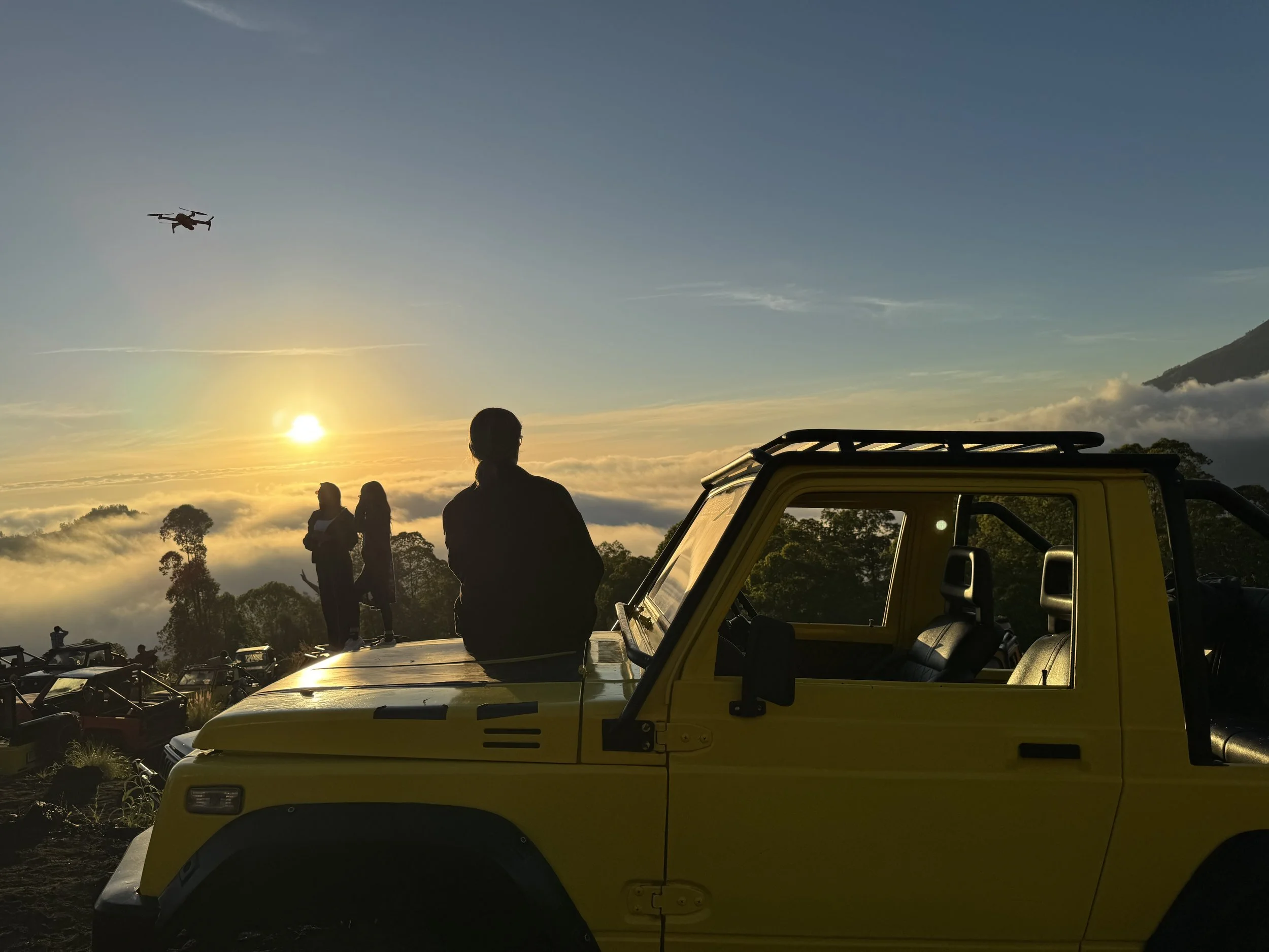 People standing near a yellow off-road vehicle and a yellow truck during a sunset or sunrise, with a drone flying in the sky above and trees and clouds in the background.