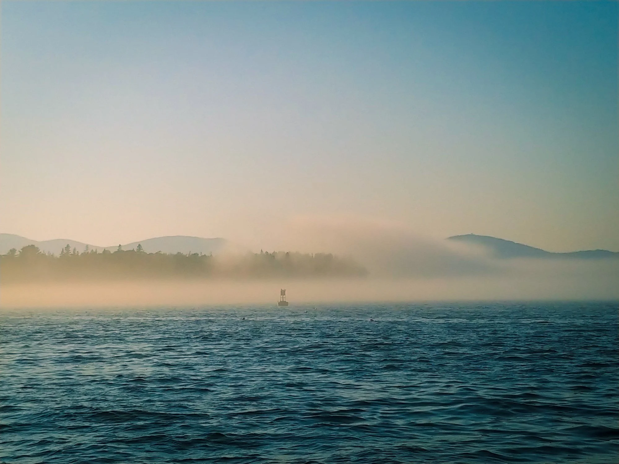 Fog bank rolling over the water just outside Manset, with dark blue waves, a small marker, and soft silhouettes of hills and shoreline in the distance.