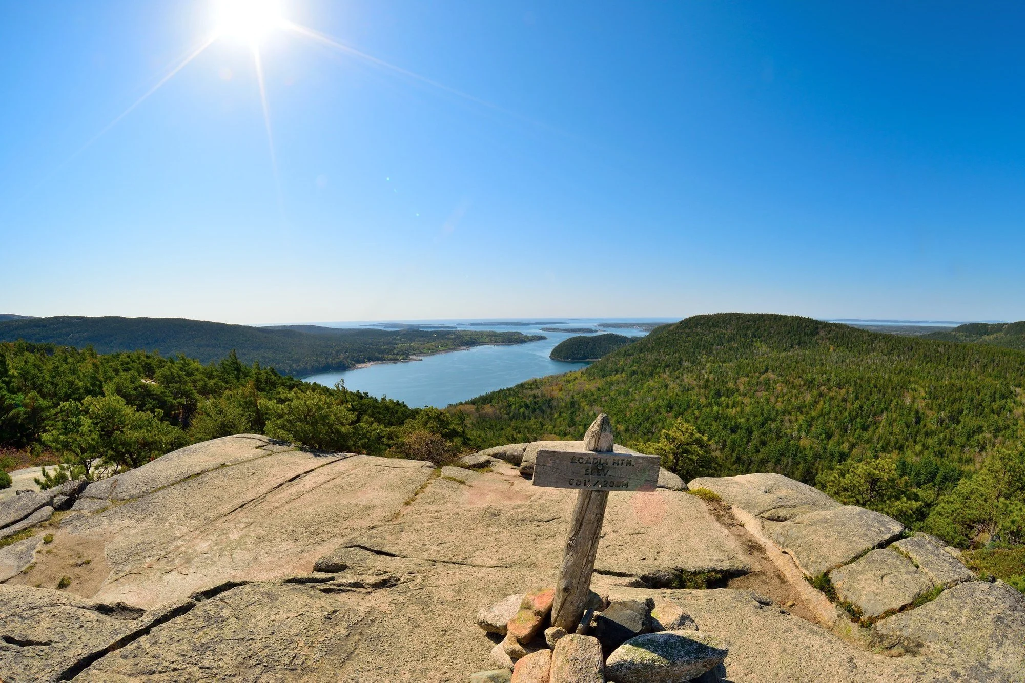 View from the summit of Acadia Mountain, with a wooden elevation sign, granite ledges, forested hills, blue water, and islands under a bright sky.