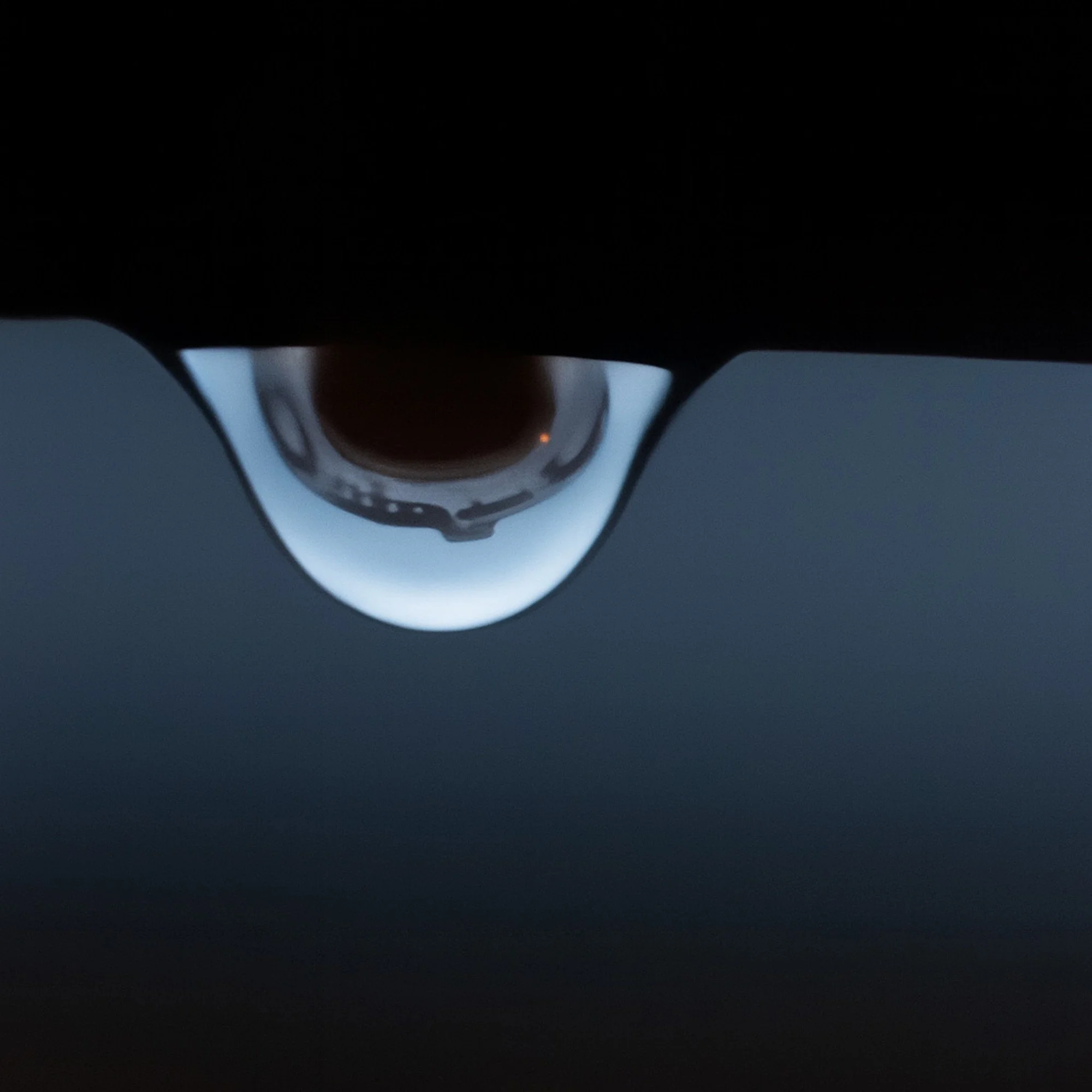 Close-up of a single raindrop hanging from the edge of a deck table, resembling an eye with an upside-down reflection of a house inside it.