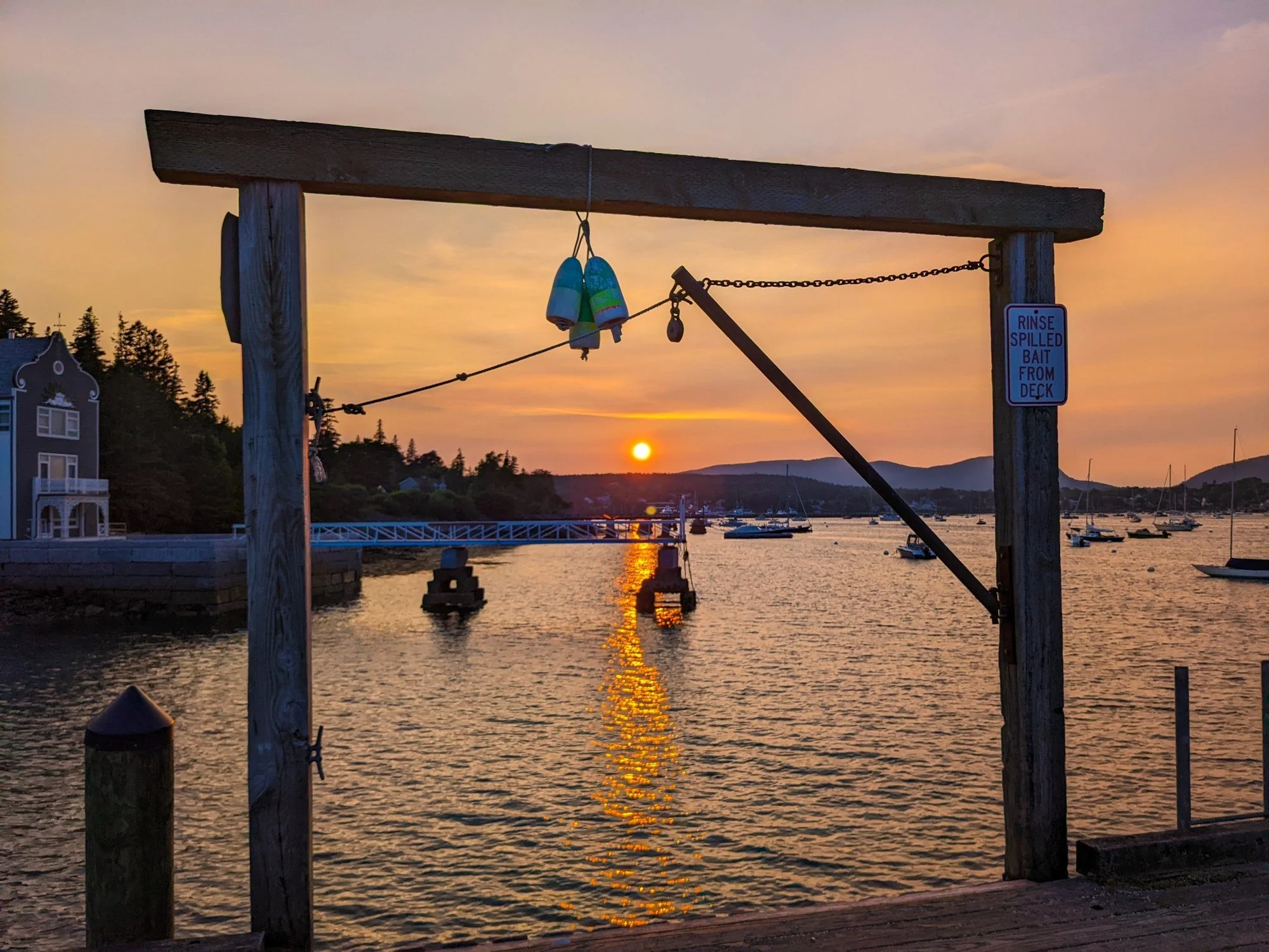 Sunset over Manset Harbor on Mount Desert Island, Maine, seen from the dock with boats, moorings, a bait hoist, and golden light on the water.