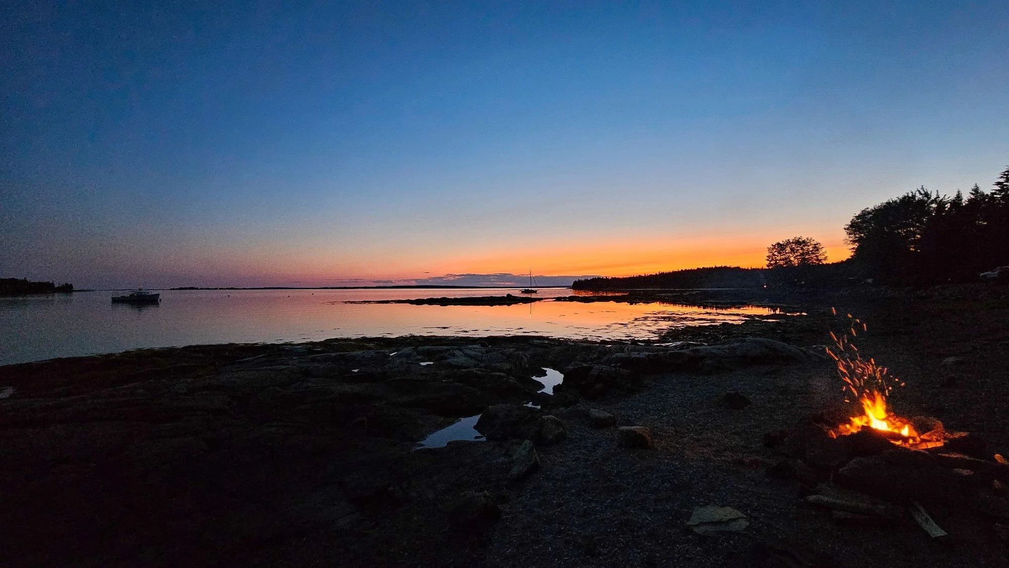 Campfire burning on the rocky shore at sunset on the Algerine Coast in Seal Cove, with boats at anchor and orange light fading over the water.