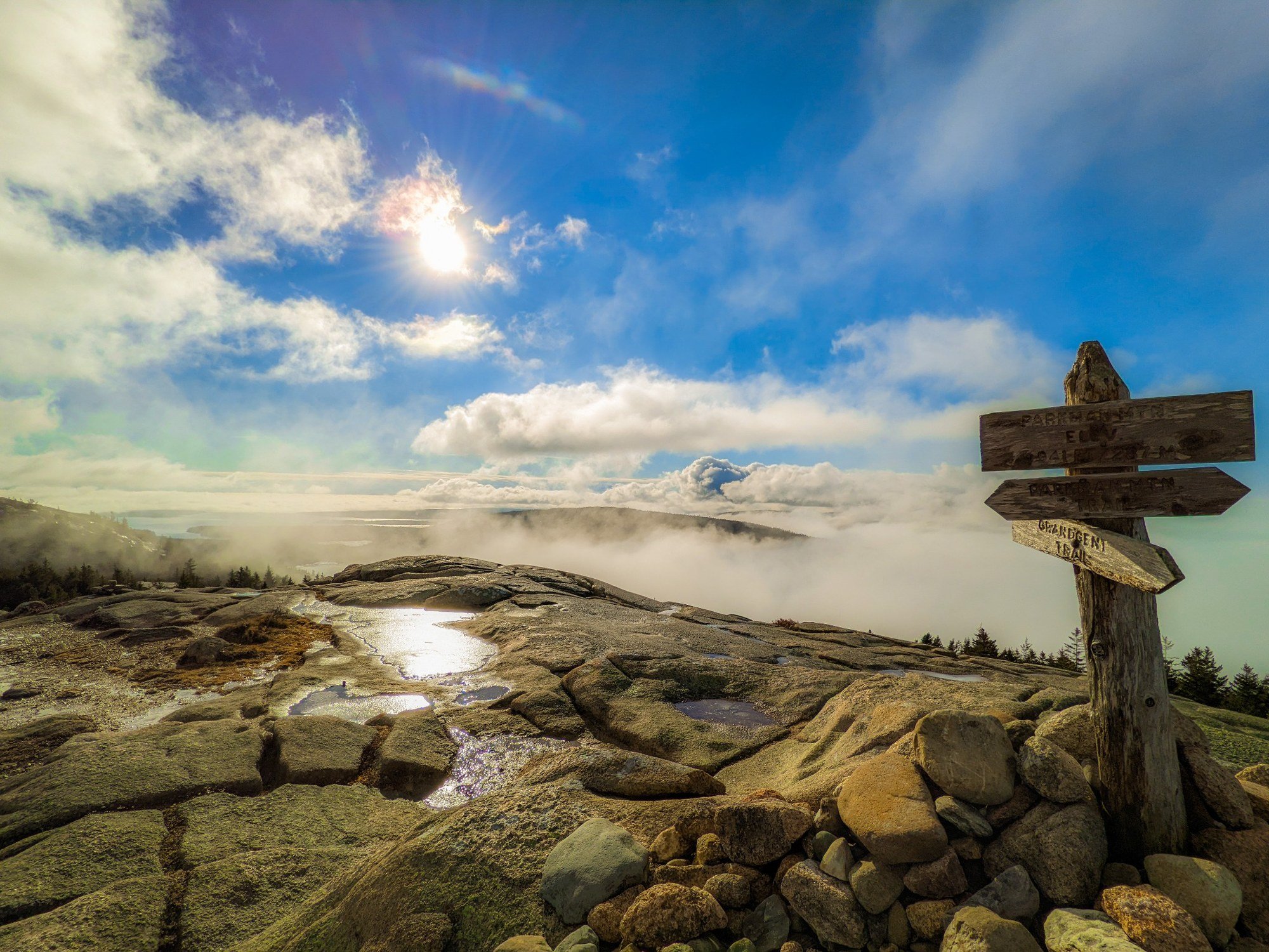 View from Parkman Mountain in Acadia National Park, with sun, clouds, fog, granite ledges, trail signs, and Mount Desert Island below.