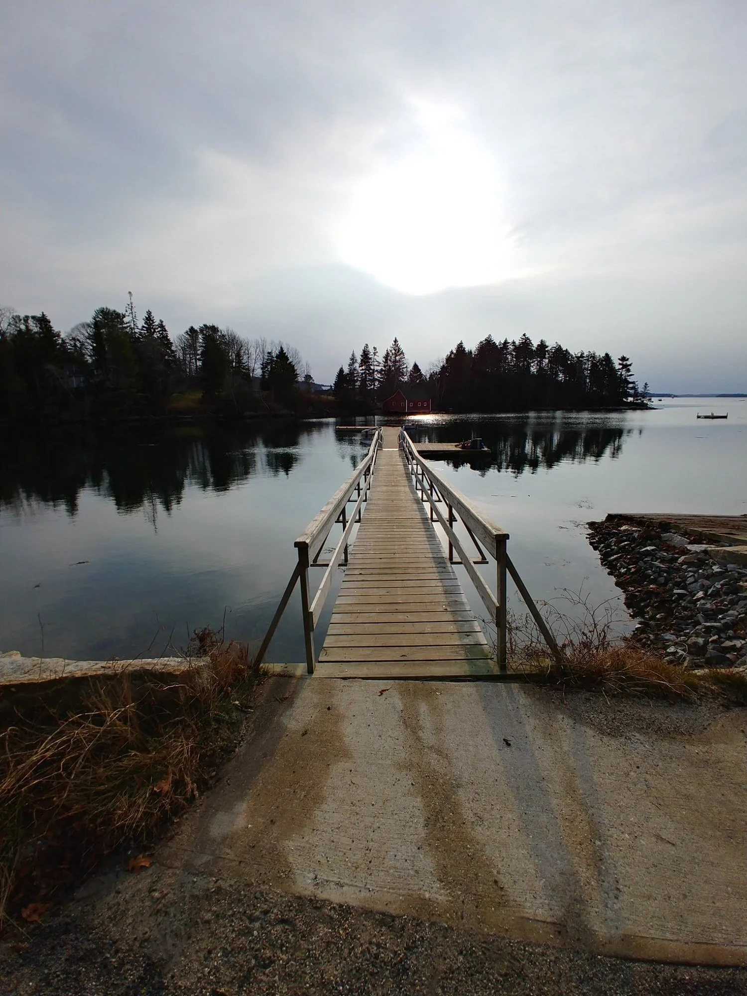 Wooden dock leading across calm water toward a small tree-covered island and red building, with winter light breaking through a bright cloudy sky