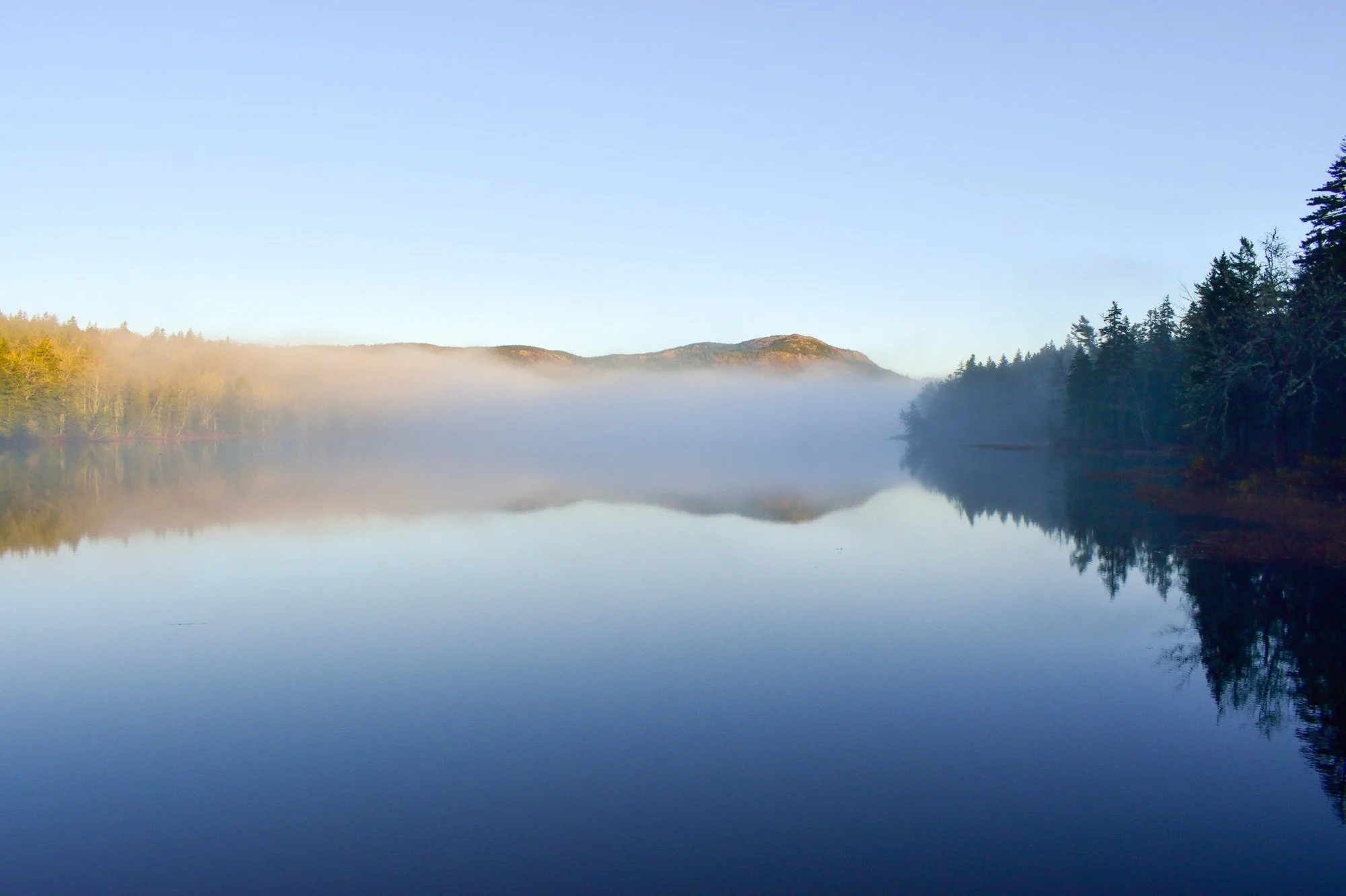 Morning fog over Little Long Pond on Mount Desert Island, Maine, with still water, reflections, forested shoreline, and hills in the distance.