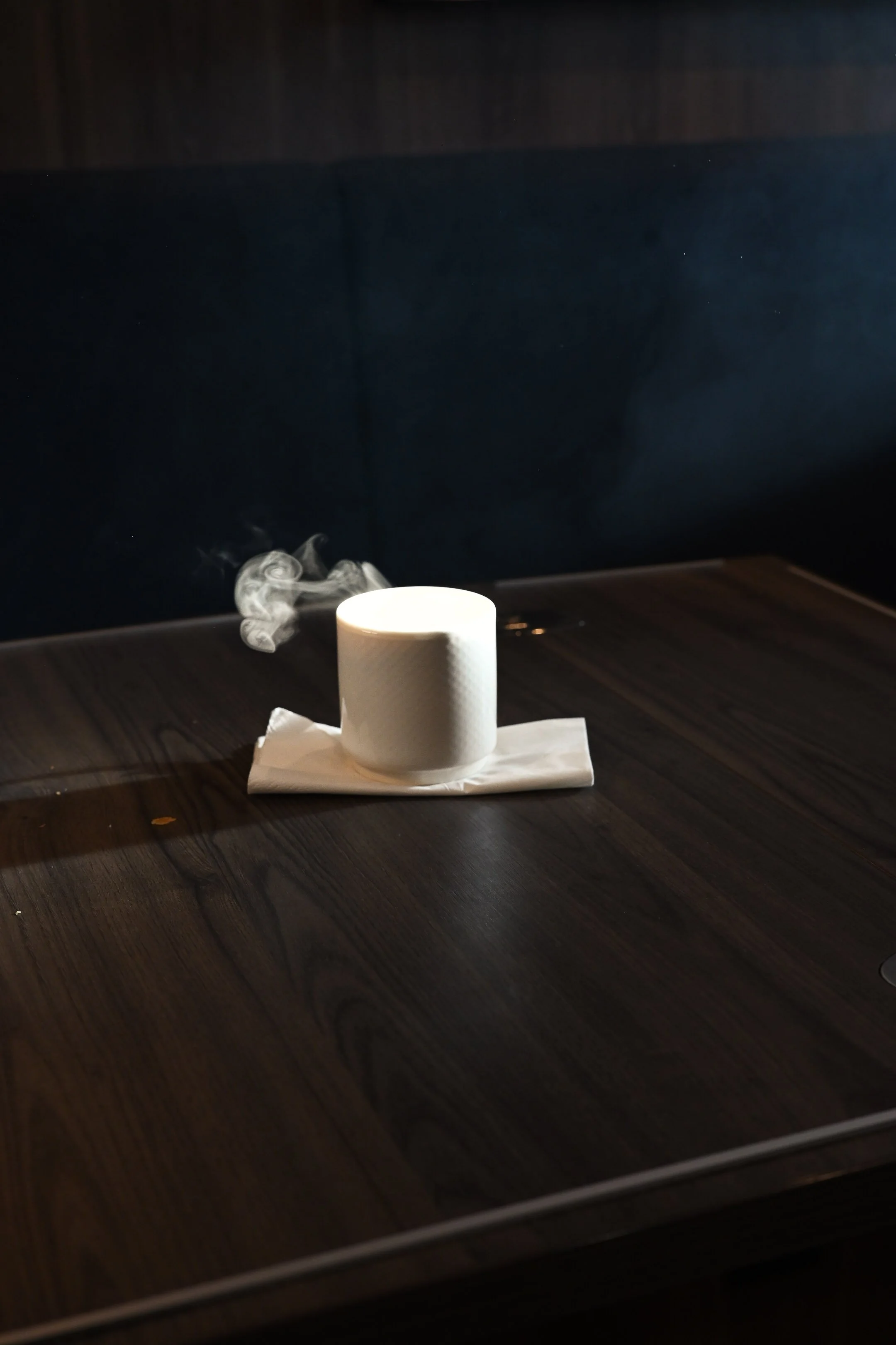 A white candle burning on a dark wooden table with a paper towel underneath, and thin wisps of smoke rising from the candle.