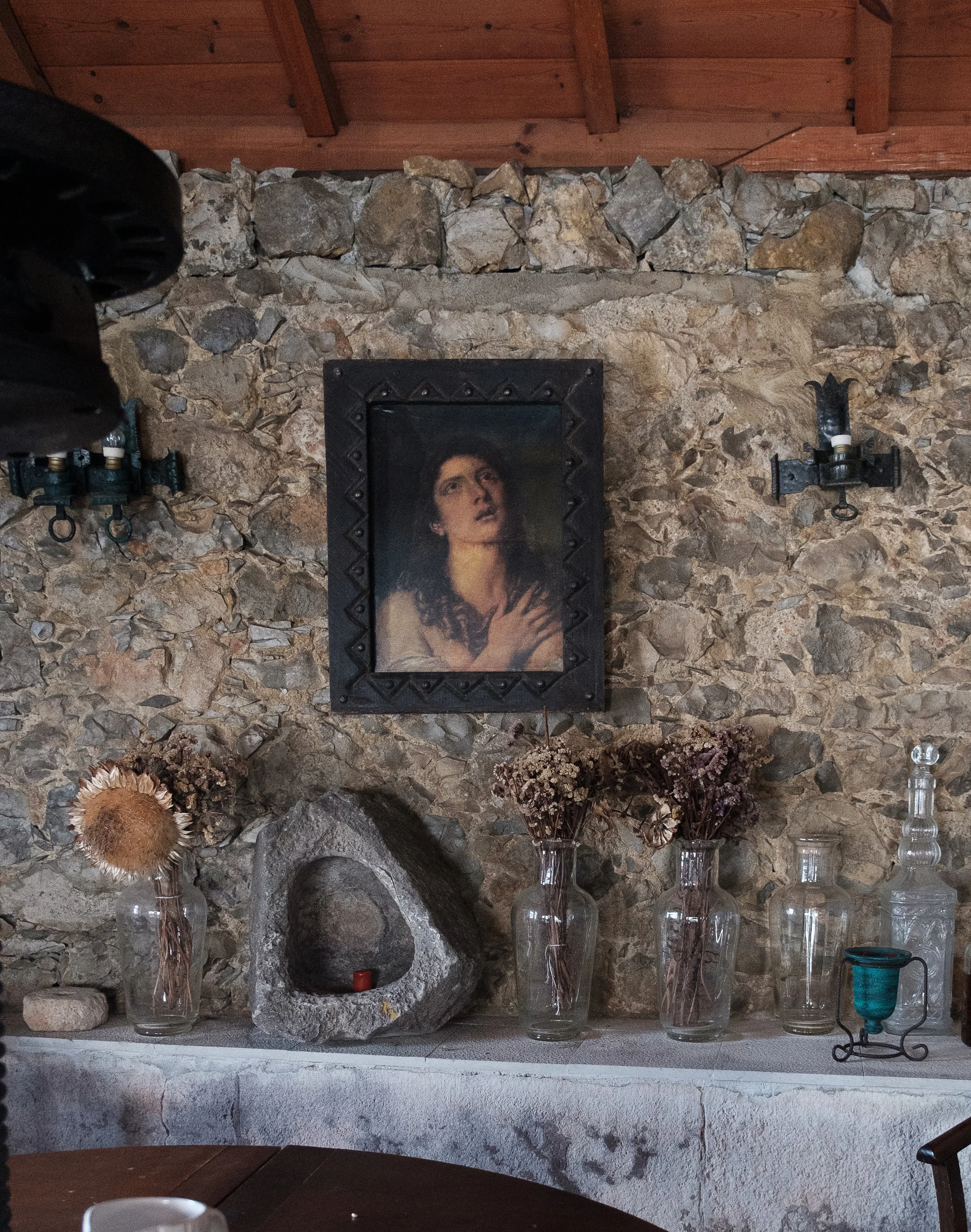 A rustic interior with a stone wall, centered portrait of a woman, and decorative dried flowers in glass vases on a shelf. There are vintage wall-mounted lamps and a wooden ceiling.