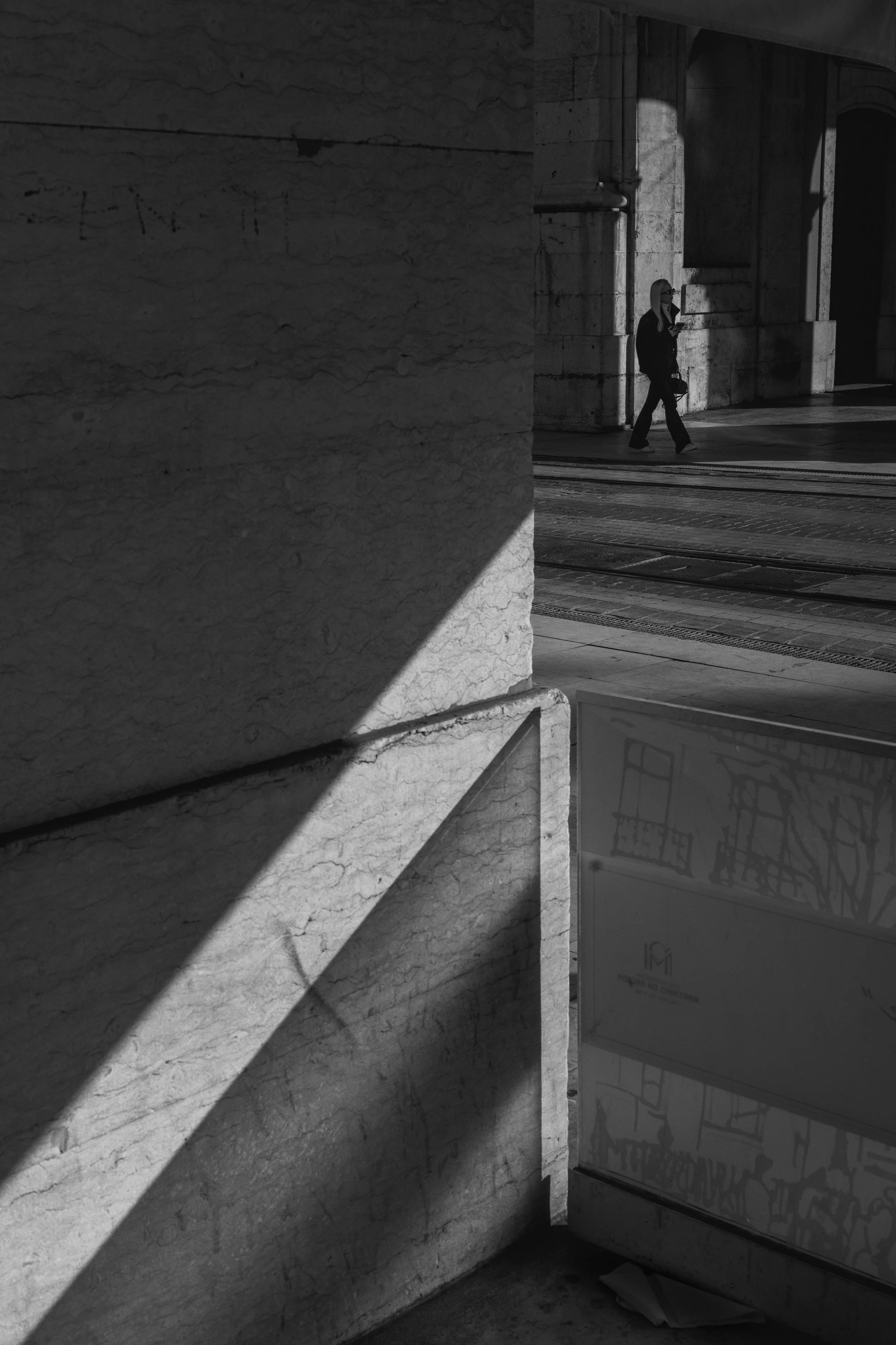 A woman with long blonde hair, wearing glasses, a dark jacket, and jeans, walking along a sidewalk while looking at her phone. She is next to large stone columns and a building with arched openings, in shadow and sunlight.