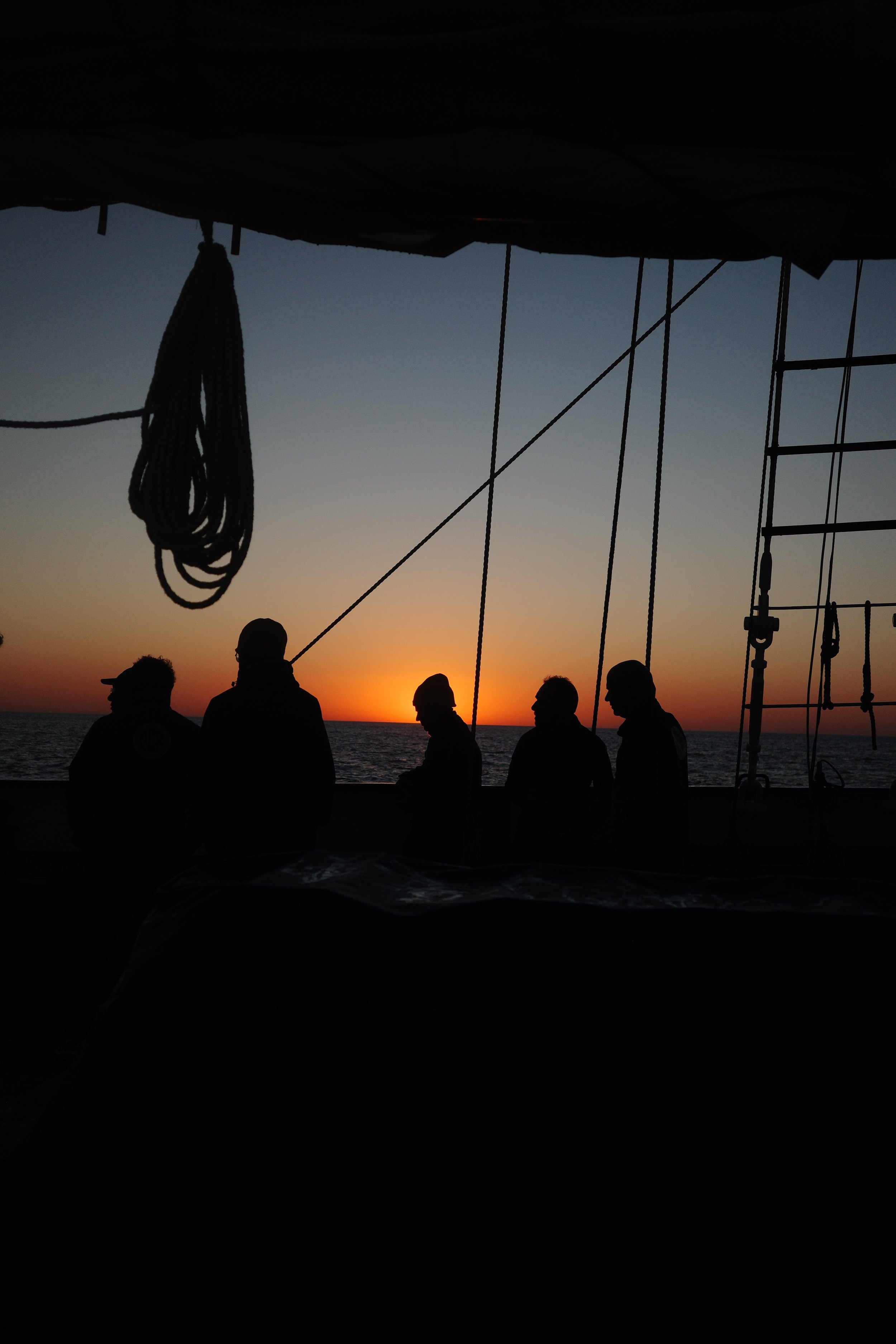 Silhouettes of people on a boat during sunset, with ropes and rigging visible in the foreground and the ocean in the background.