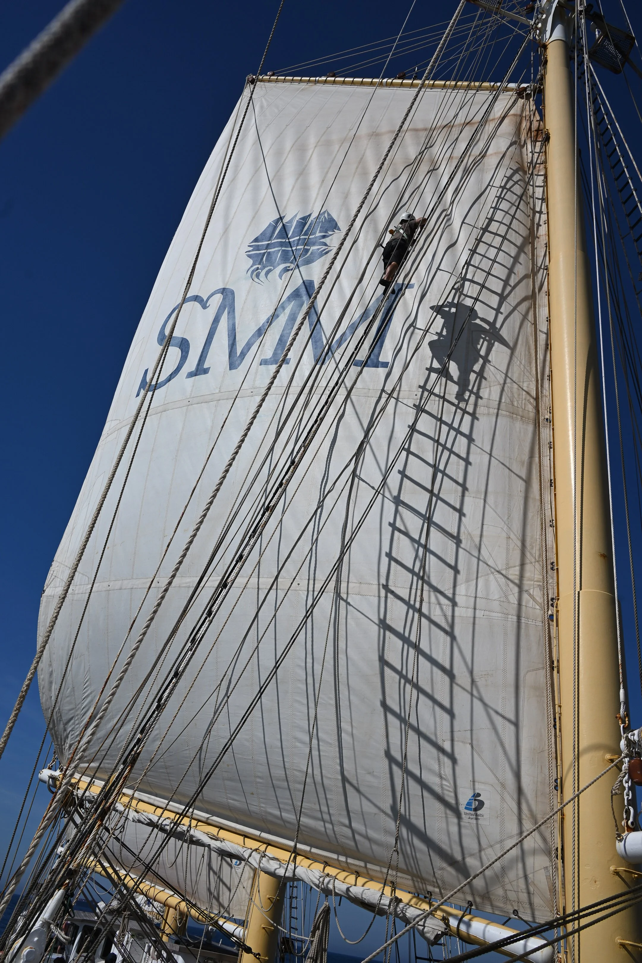 A person climbing the rigging of a tall ship with sails and ropes against a blue sky.