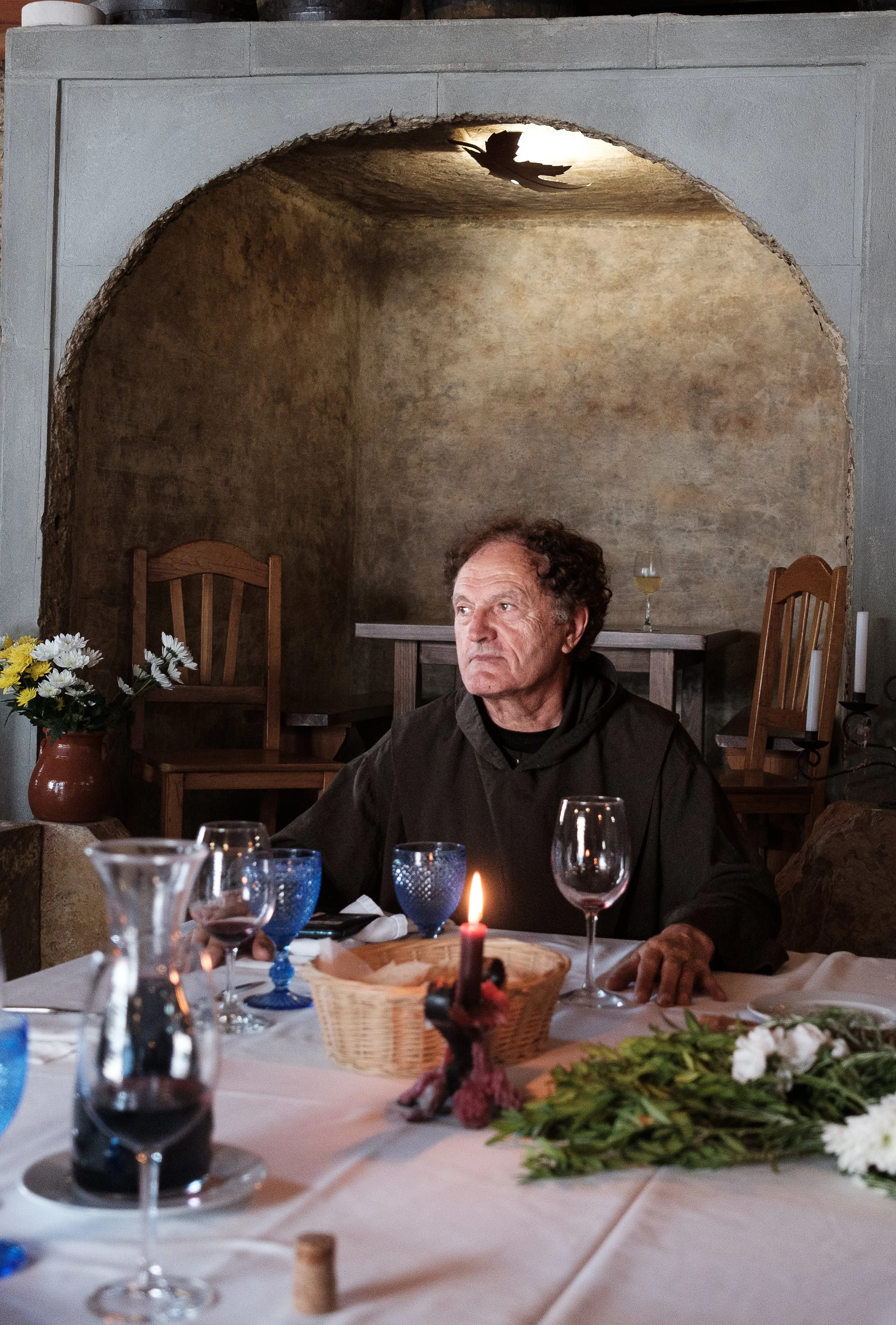A man with curly hair and a black jacket sitting at a dinner table with wine glasses, a candle, and a bouquet of white flowers in a cozy room with a rustic stone wall and wooden chairs in the background.