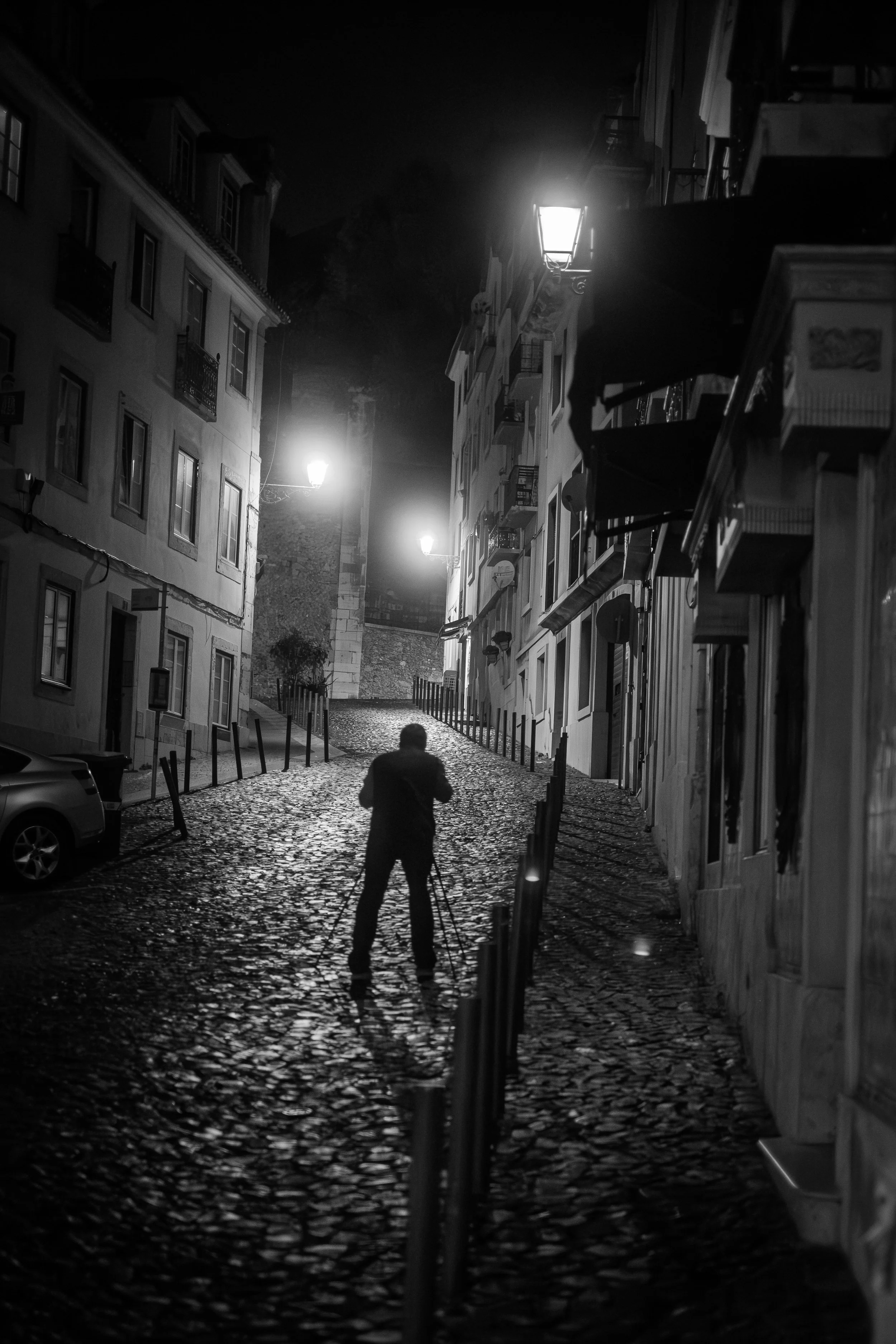 A person standing on a cobblestone street at night, holding a tripod, with lit street lamps illuminating the scene. Buildings line the street, some with balconies and signage, and there is a car parked on the left side.