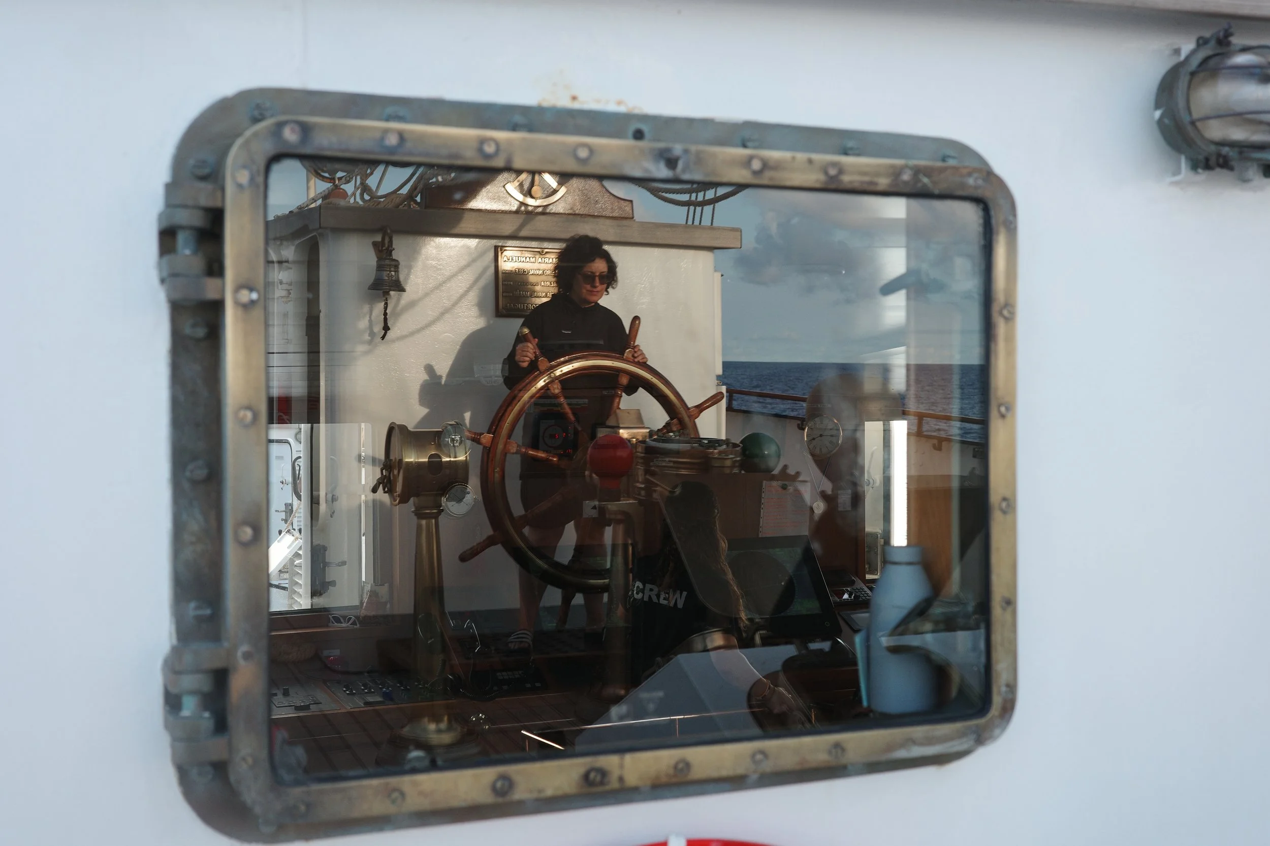 Reflection of a woman at a ship's bridge window, steering wheel, dashboard, and gauge visible inside the ship