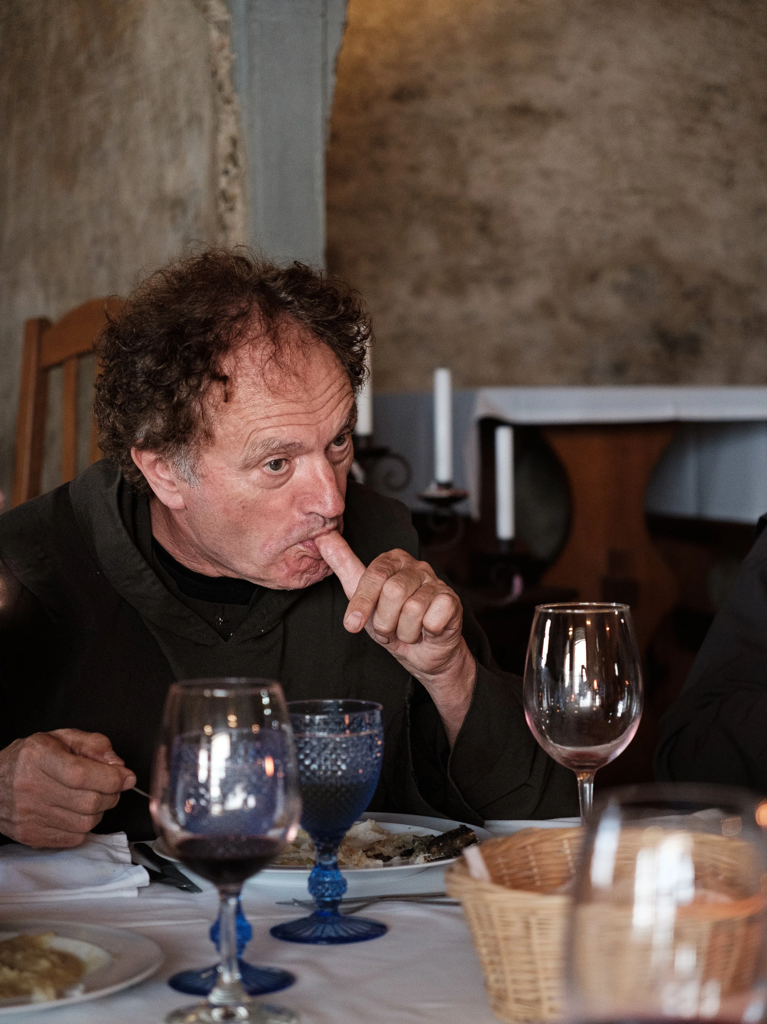 A man with curly hair is sitting at a dining table, placing his finger on his lips. The table has several wine glasses, a basket, and plates of food. The background features a stone wall and a piece of furniture.