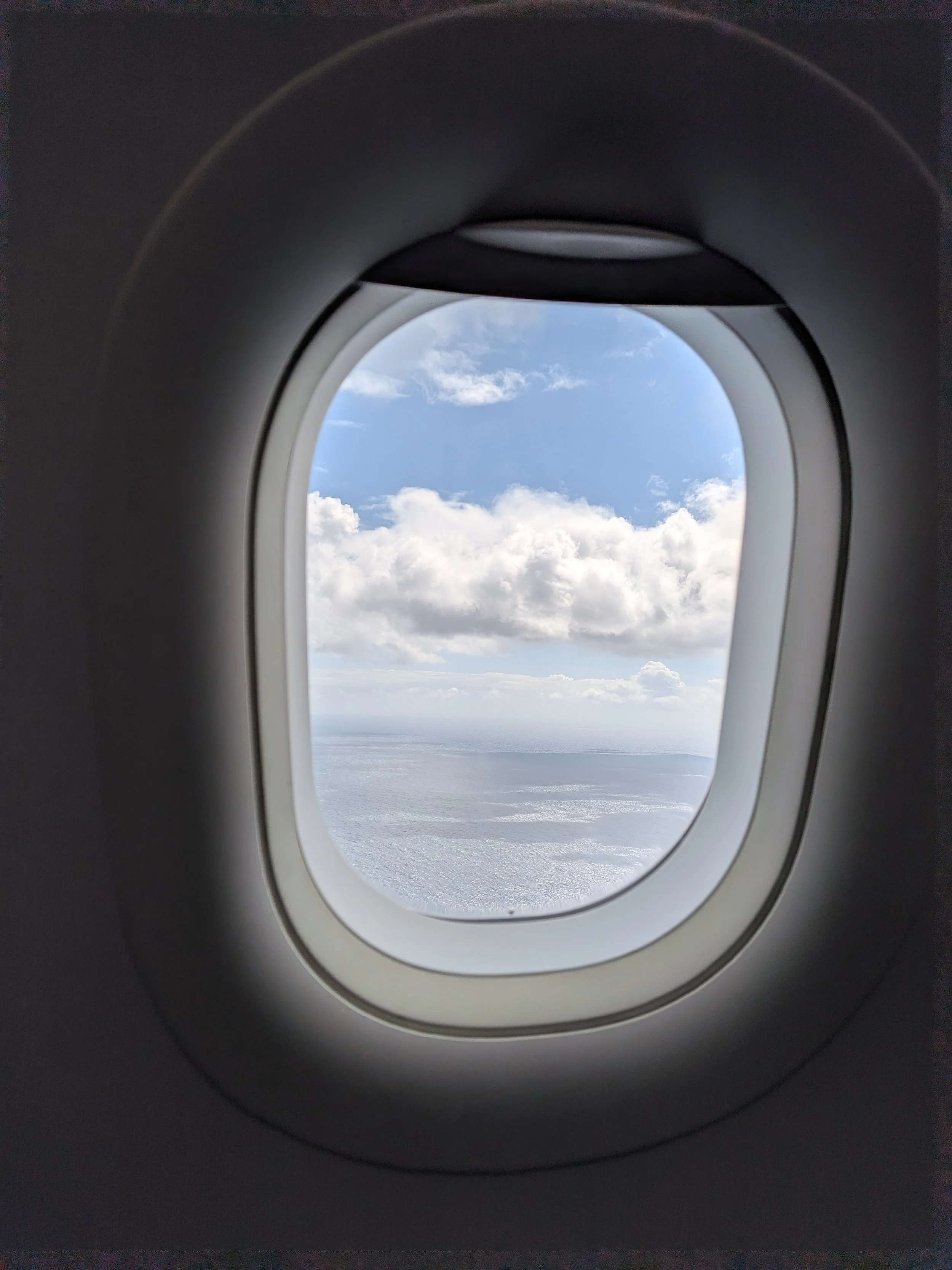 View through an airplane window showing clouds and blue sky outside.